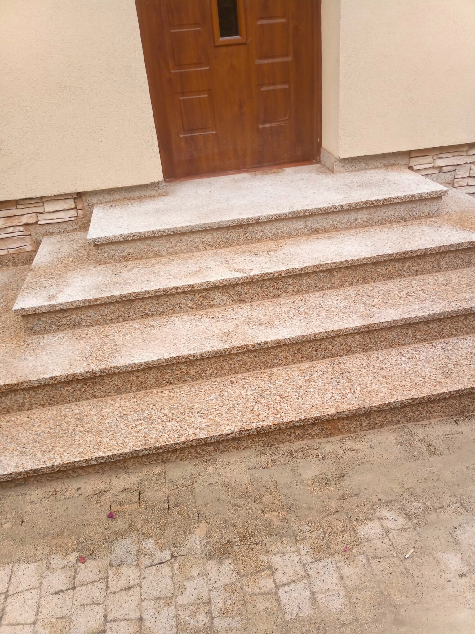 Zewnętrzne schody wejściowe wyłożone beżowym kamieniem z brązowymi i białymi drobinkami, prowadzące do drewnianych drzwi wejściowych z szybką, na tle beżowej elewacji budynku z elementami kamiennej...