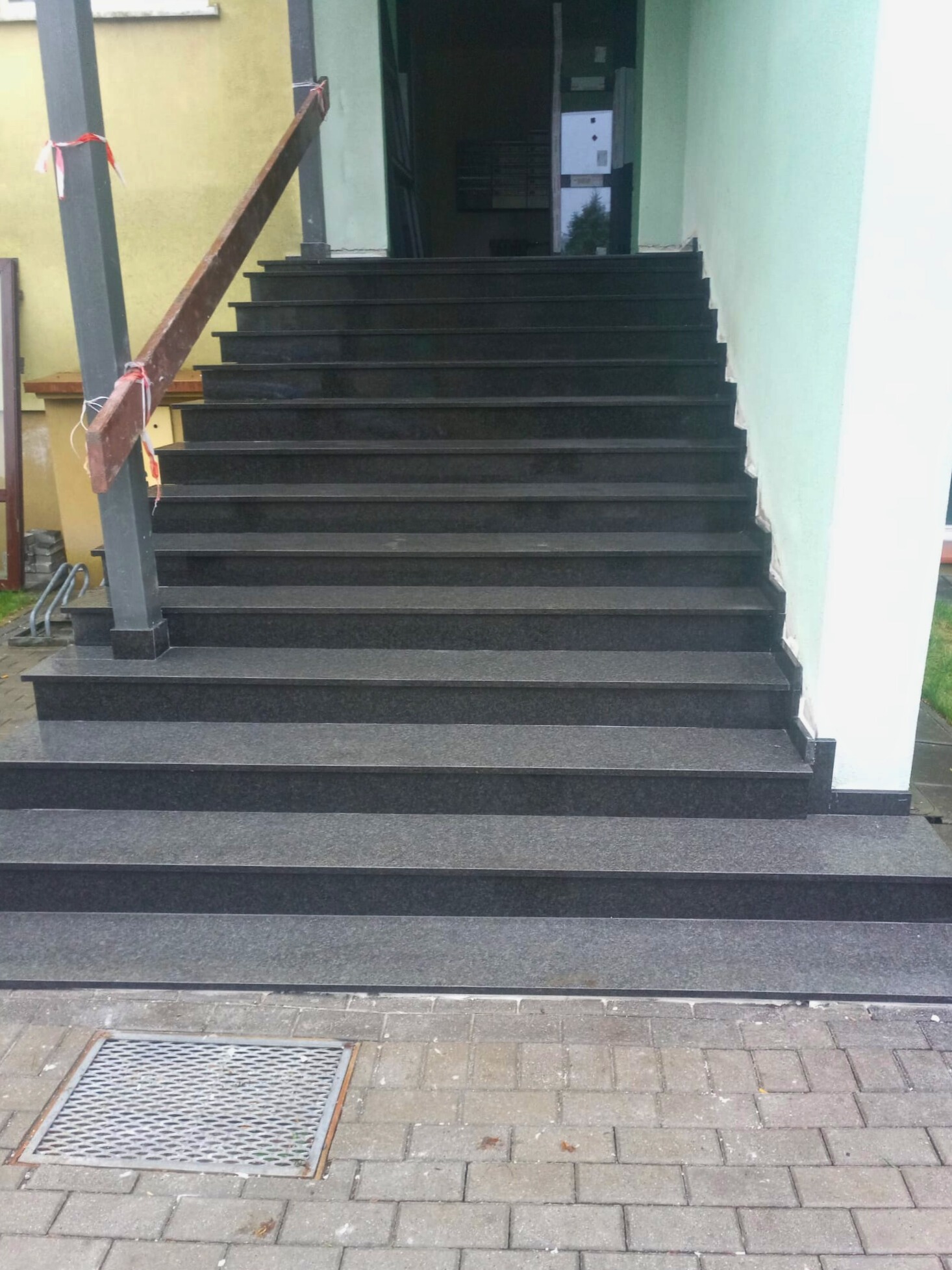 Zewnętrzne schody wejściowe wyłożone szarym kamieniem, z widocznym fragmentem elewacji budynku w kolorze jasnozielonym oraz żółtym, zabezpieczone drewnianą barierką tymczasową.