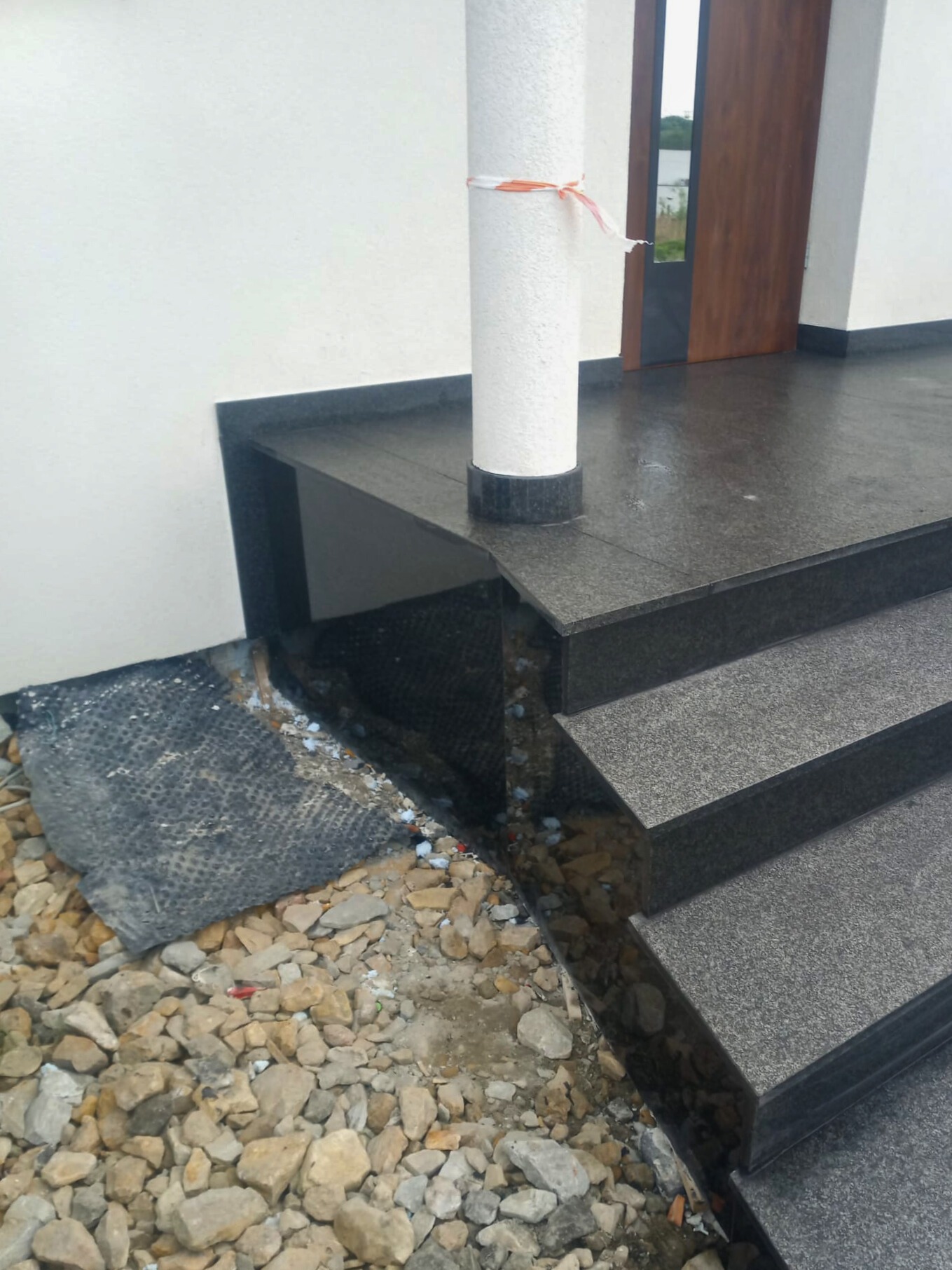 Granitowe schody wejściowe do domu w trakcie montażu, widoczne kamienie, geowłóknina i kolumna z taśmą zabezpieczającą.
