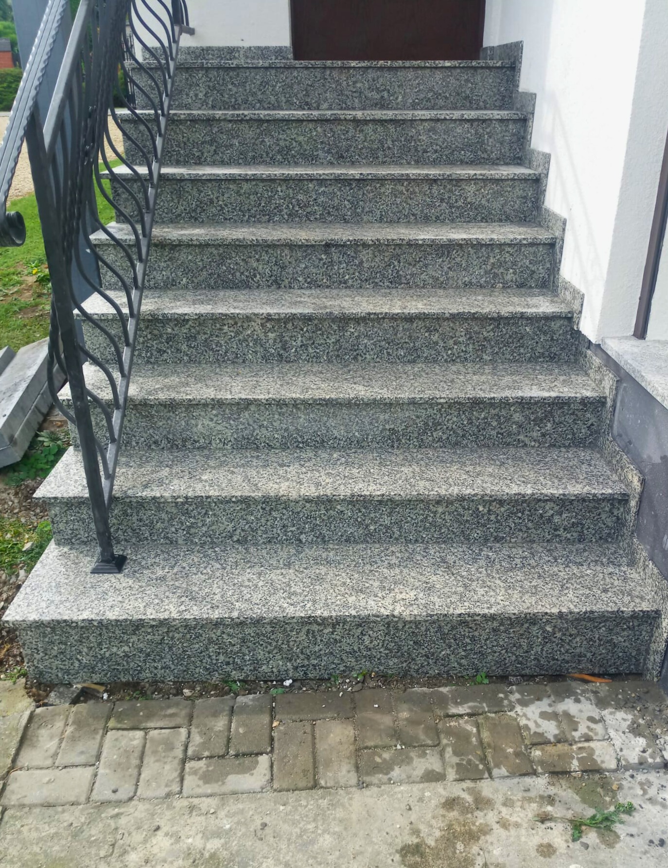 Zewnętrzne schody wejściowe wyłożone szarym, nakrapianym granitem, z czarną, kutą balustradą, prowadzące do brązowych drzwi.