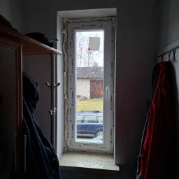 SKO-MONT OKNA - Wnętrze z nowym, białym oknem PCV Aluplast po montażu, widoczne pianka montażowa, szyba z naklejką, za oknem budynek gospodarczy i zaparkowany samochód.