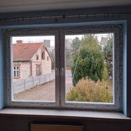 SKO-MONT OKNA - Dwukomorowe okno PCV po montażu, widok z pianką montażową. Za oknem dom z cegły i drzewa. Drobne zabrudzenia na parapecie. Szare ściany wewnątrz pomieszczenia.