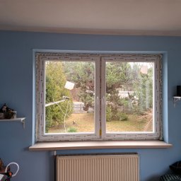 SKO-MONT OKNA - Nowe, dwuskrzydłowe okno PCV z pianką montażową, widok na ogród. Świeżo zamontowane, z widocznymi etykietami producenta na ramie. Błękitne ściany w tle.