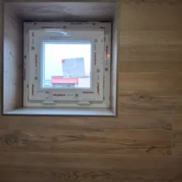 Białe okno PCV marki Aluplast, wbudowane w ścianę wyłożoną panelami imitującymi drewno, widok z bliska, z naklejkami informacyjnymi na szybie.