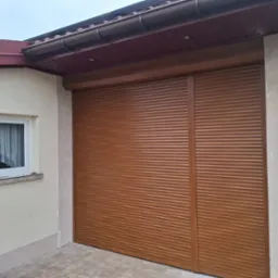Zewnętrzna, brązowa roleta garażowa zamontowana na budynku z oknem. Widoczny fragment dachu z rynną. Elewacja budynku w jasnym kolorze.