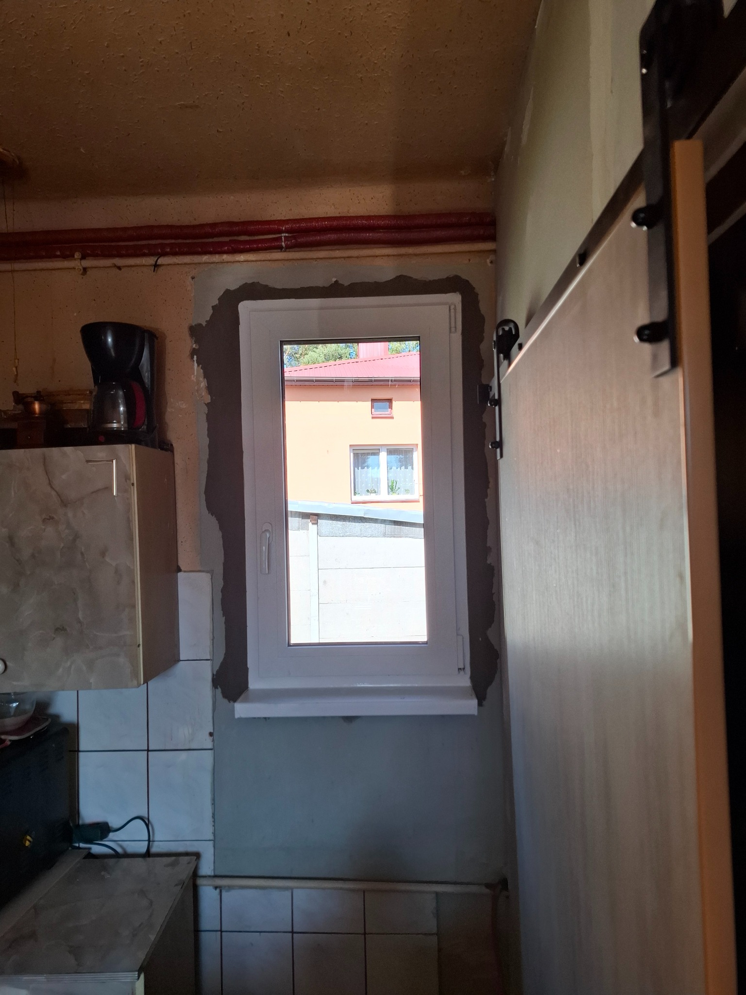 Montaż okna PCV w kuchni: biała rama okienna osadzona w surowym murze, widok na budynek z czerwonym dachem. Obok widoczna przesuwna konstrukcja drzwi.