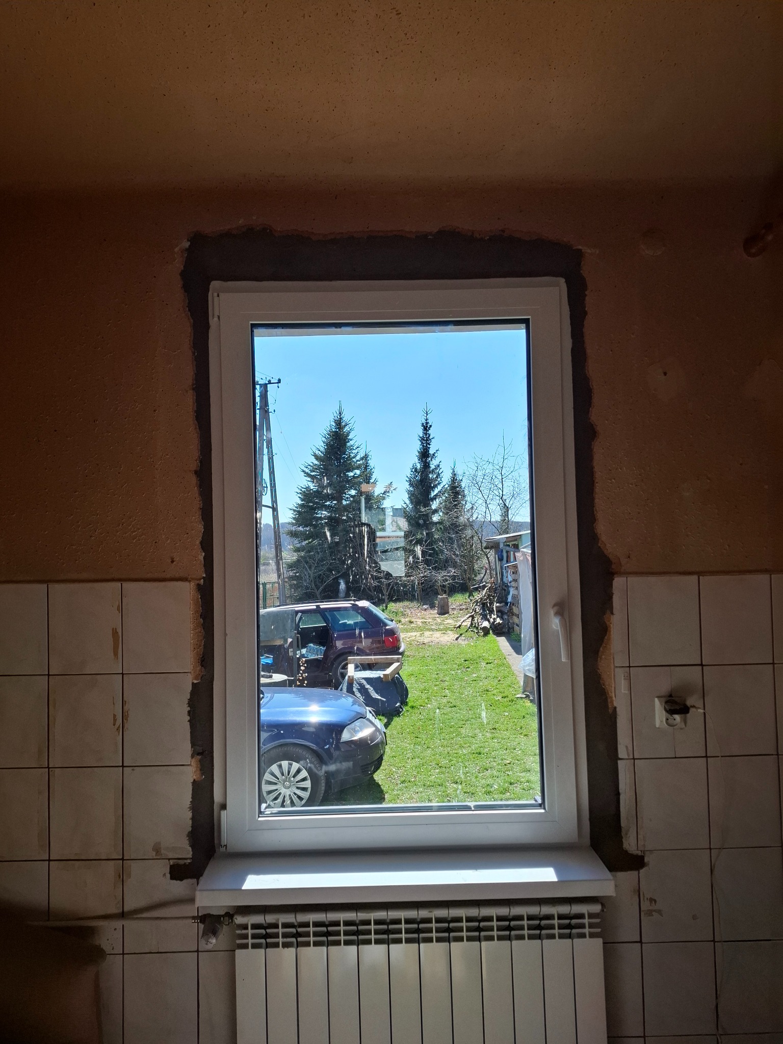 Białe okno PCV osadzone w surowym otworze, widok na podwórko z autami i zielenią. Wnętrze w trakcie remontu, z fragmentami płytek na ścianach i grzejnikiem pod oknem.