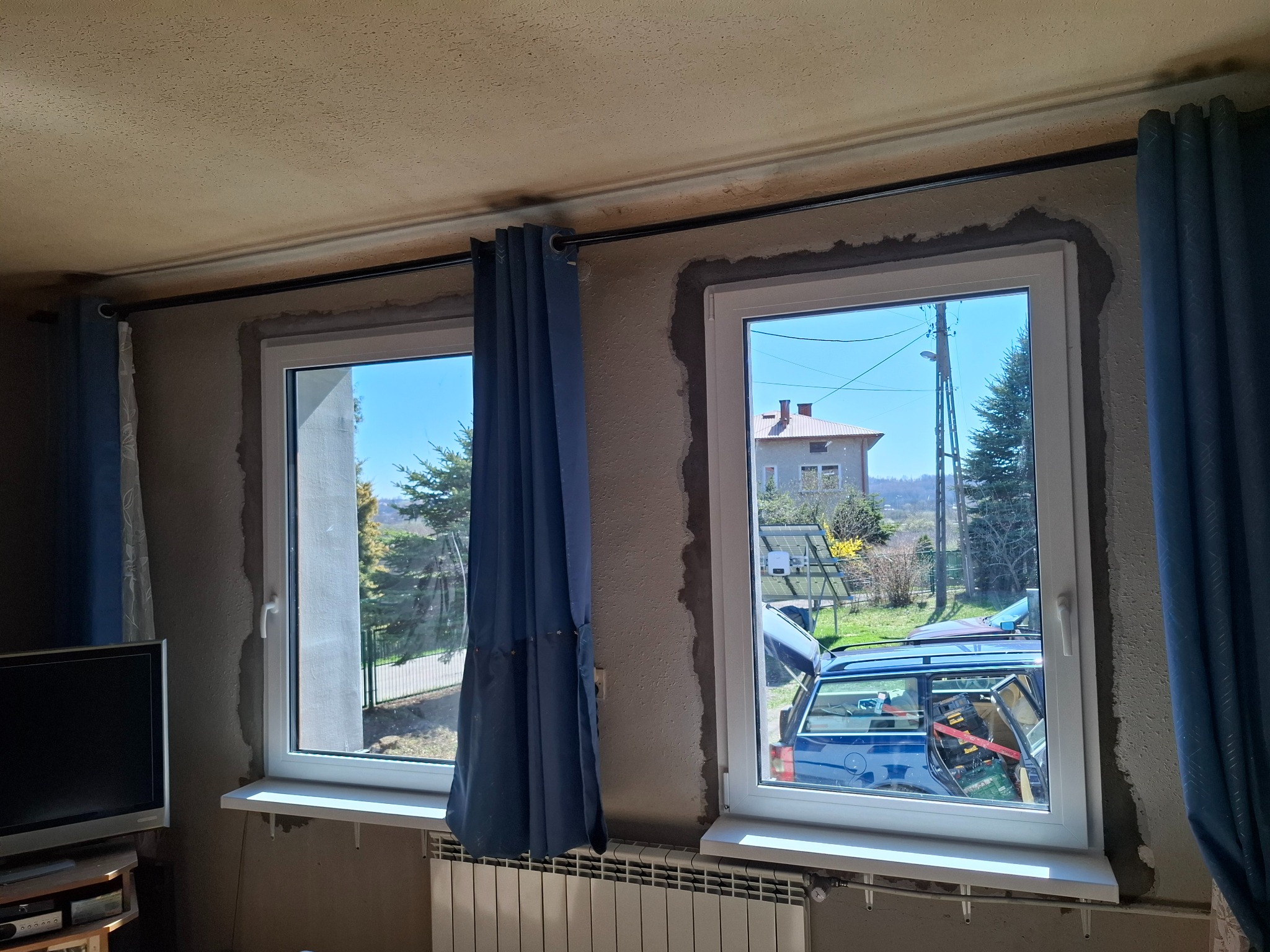 Dwa białe okna PCV po montażu, z widocznymi śladami prac murarskich na ścianie. Za oknem widok na podwórko z samochodem wypełnionym narzędziami.
