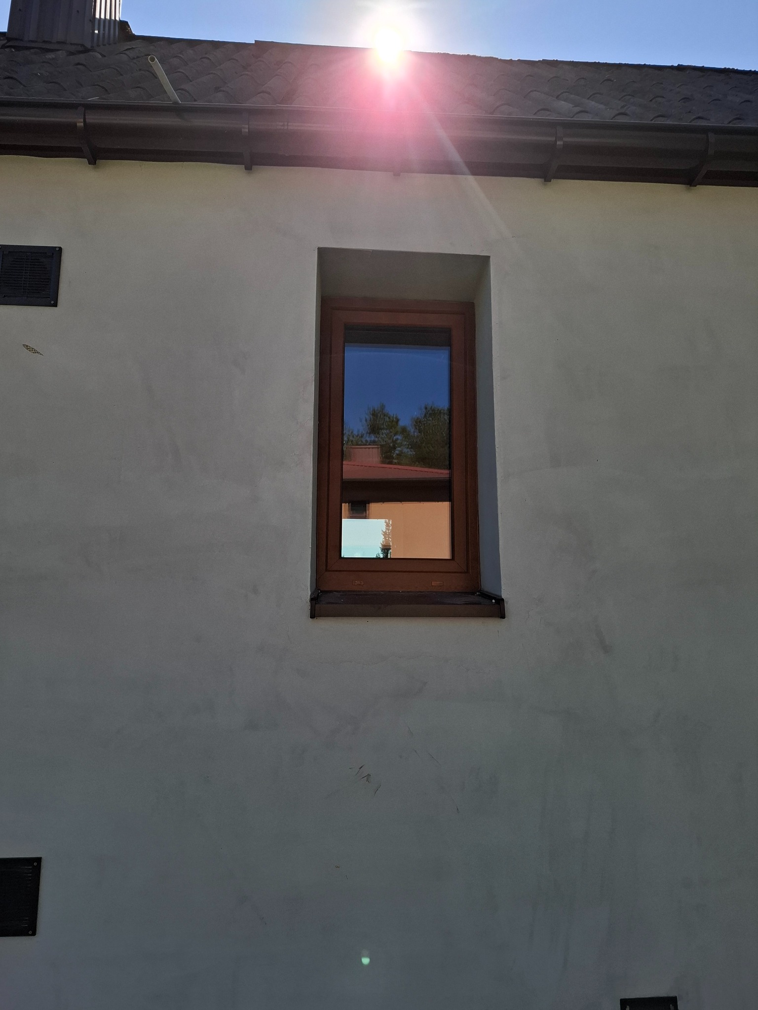 Brązowe okno w szarej ścianie domu, odbijające niebo i fragment dachu sąsiedniego budynku. Słońce w górnym rogu kadru.