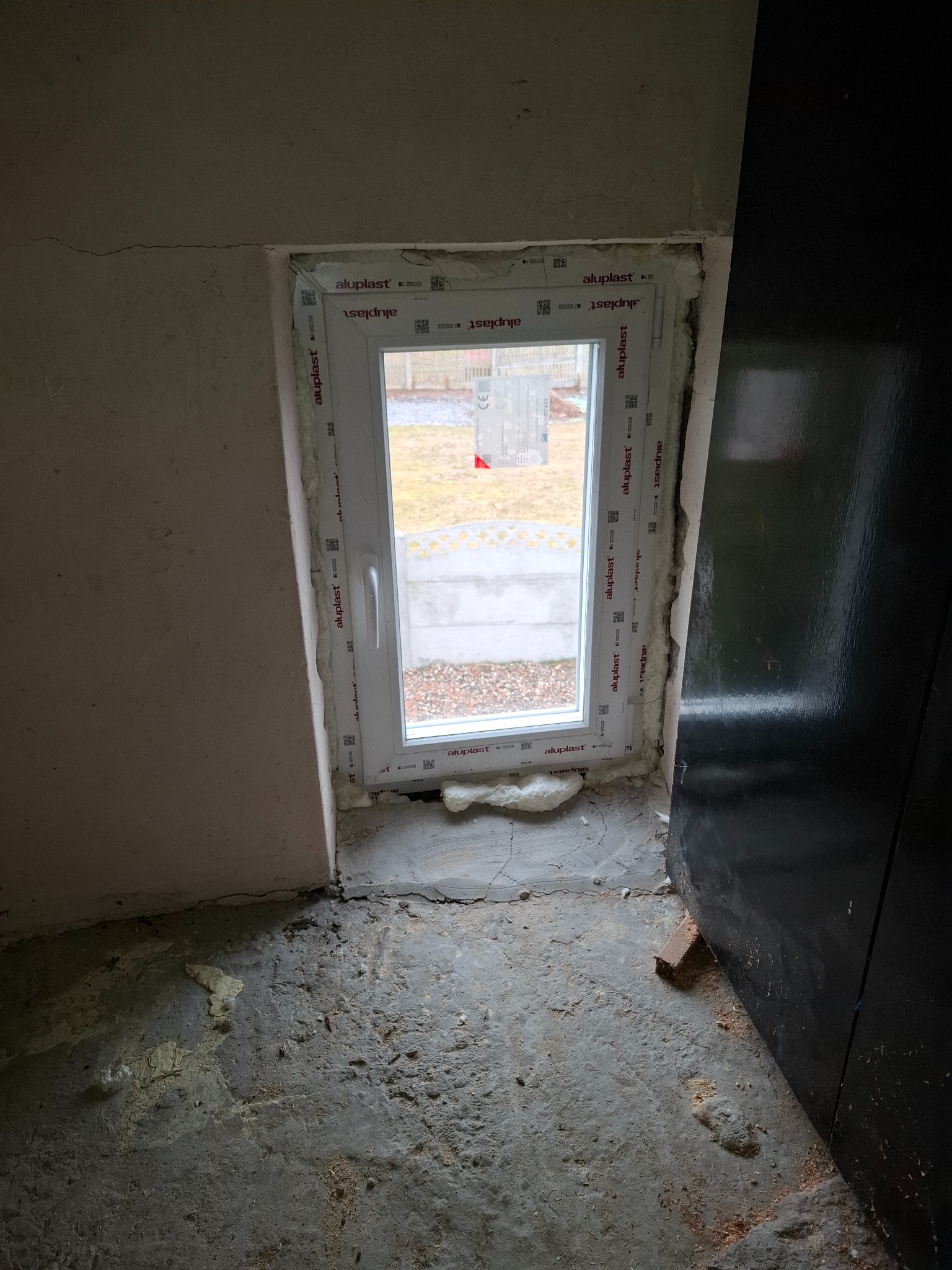 Nowe okno PCV Aluplast w surowym otoczeniu: widok z bliska na świeżo zamontowane okno, z pianką montażową i widocznym otoczeniem w stanie wykończeniowym.