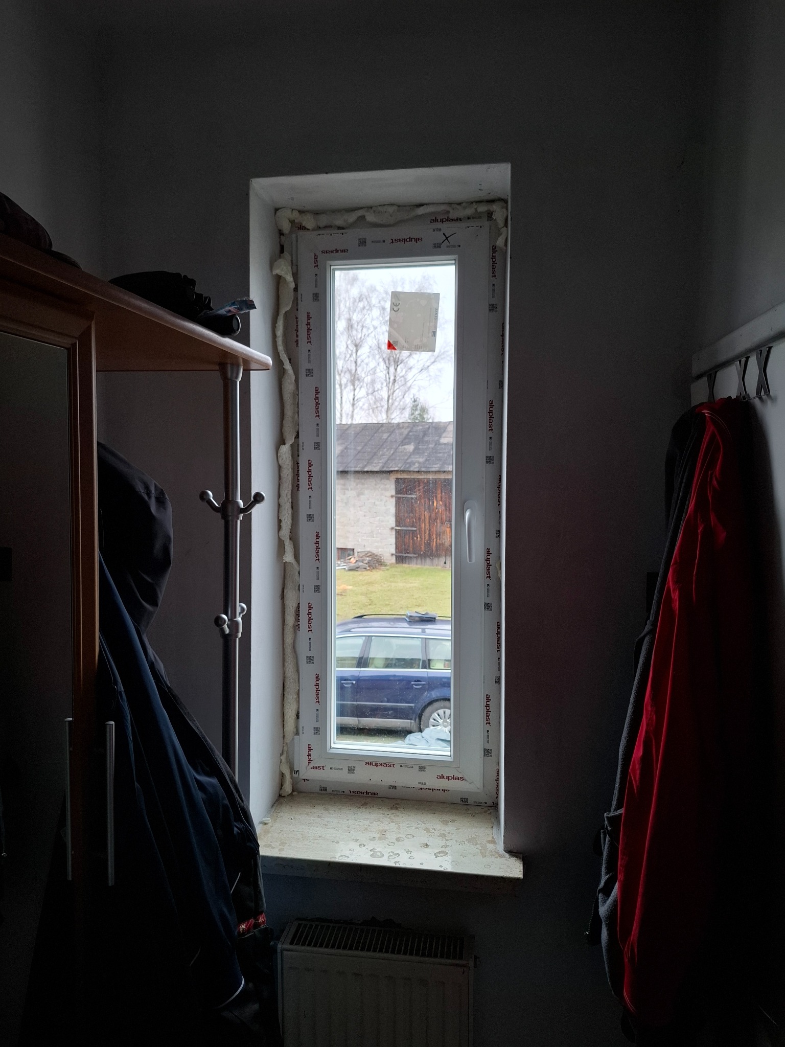 Wnętrze z nowym, białym oknem PCV Aluplast po montażu, widoczne pianka montażowa, szyba z naklejką, za oknem budynek gospodarczy i zaparkowany samochód.