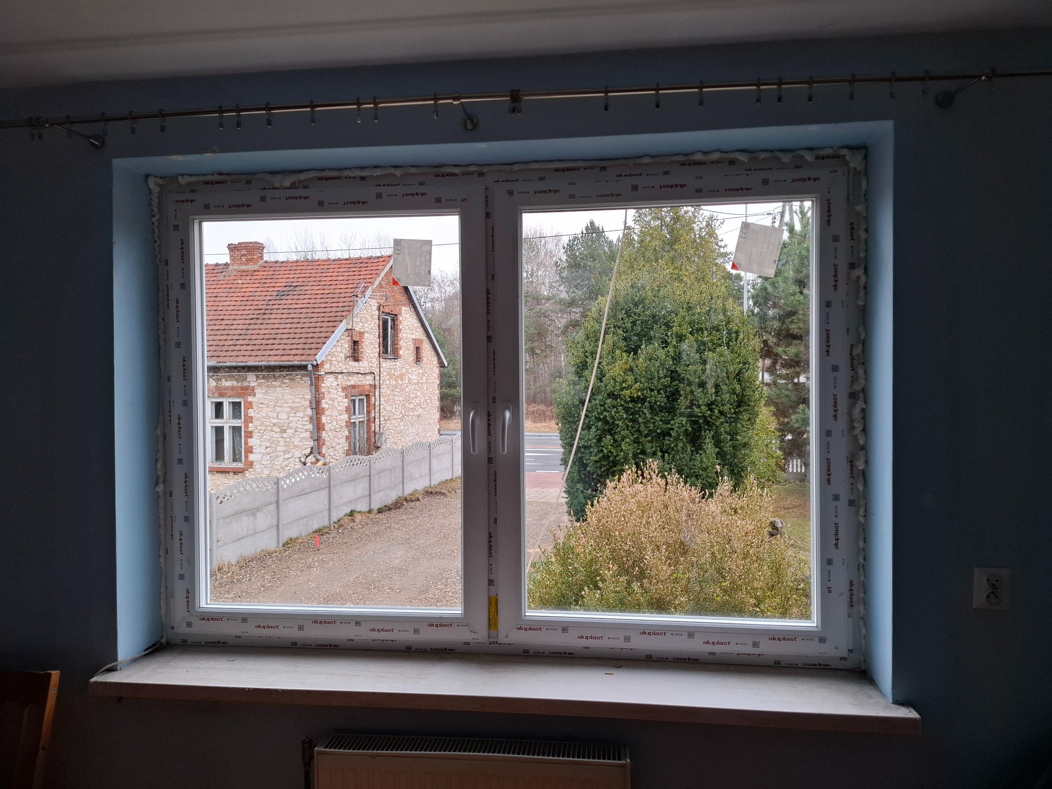 Dwukomorowe okno PCV po montażu, widok z pianką montażową. Za oknem dom z cegły i drzewa. Drobne zabrudzenia na parapecie. Szare ściany wewnątrz pomieszczenia.