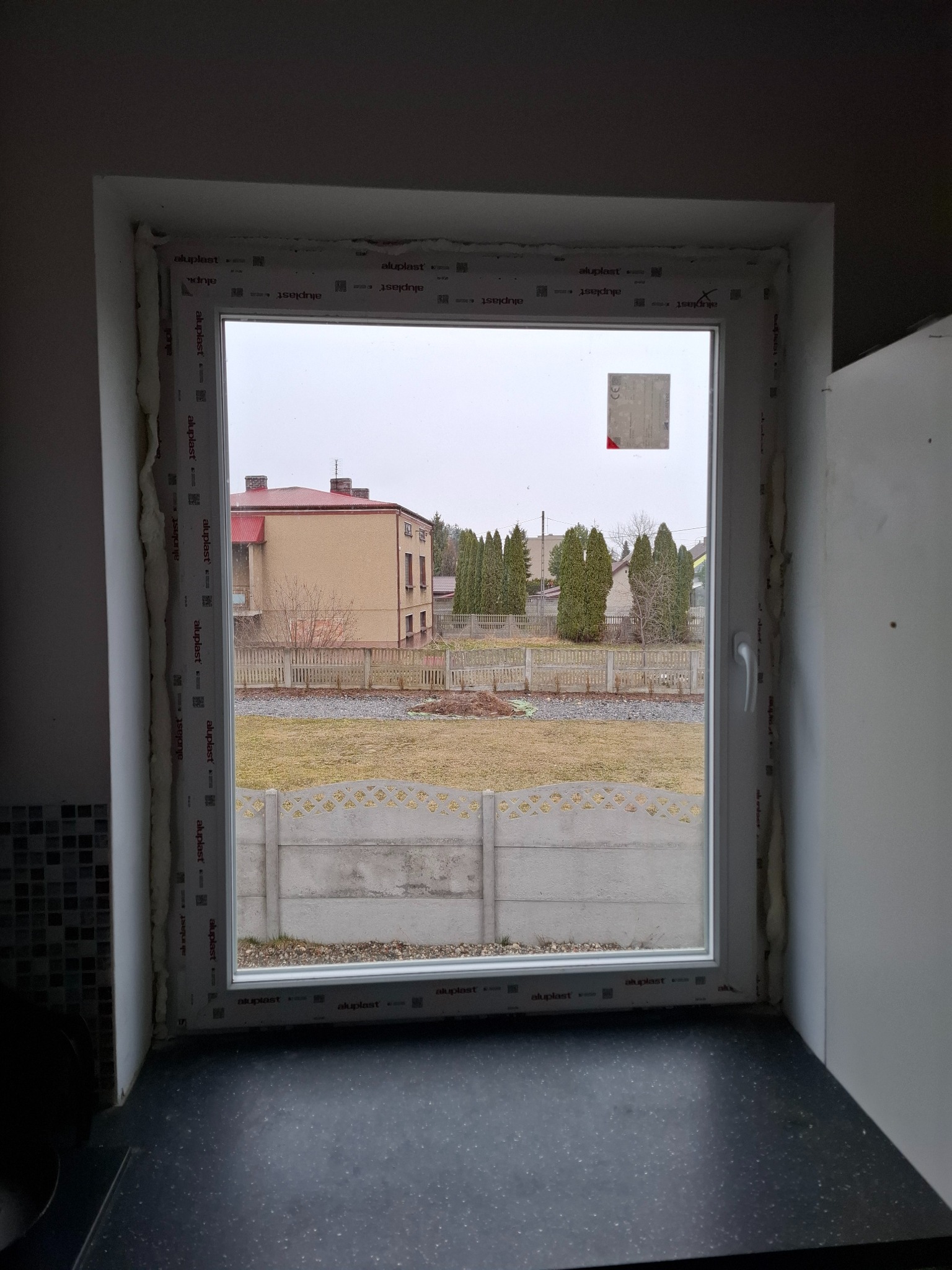 Montaż okna PCV w Bystrzanowicach. Widok na zewnątrz przez świeżo osadzone okno z pianką montażową. Nowa stolarka okienna z widokiem na ogród i dom.