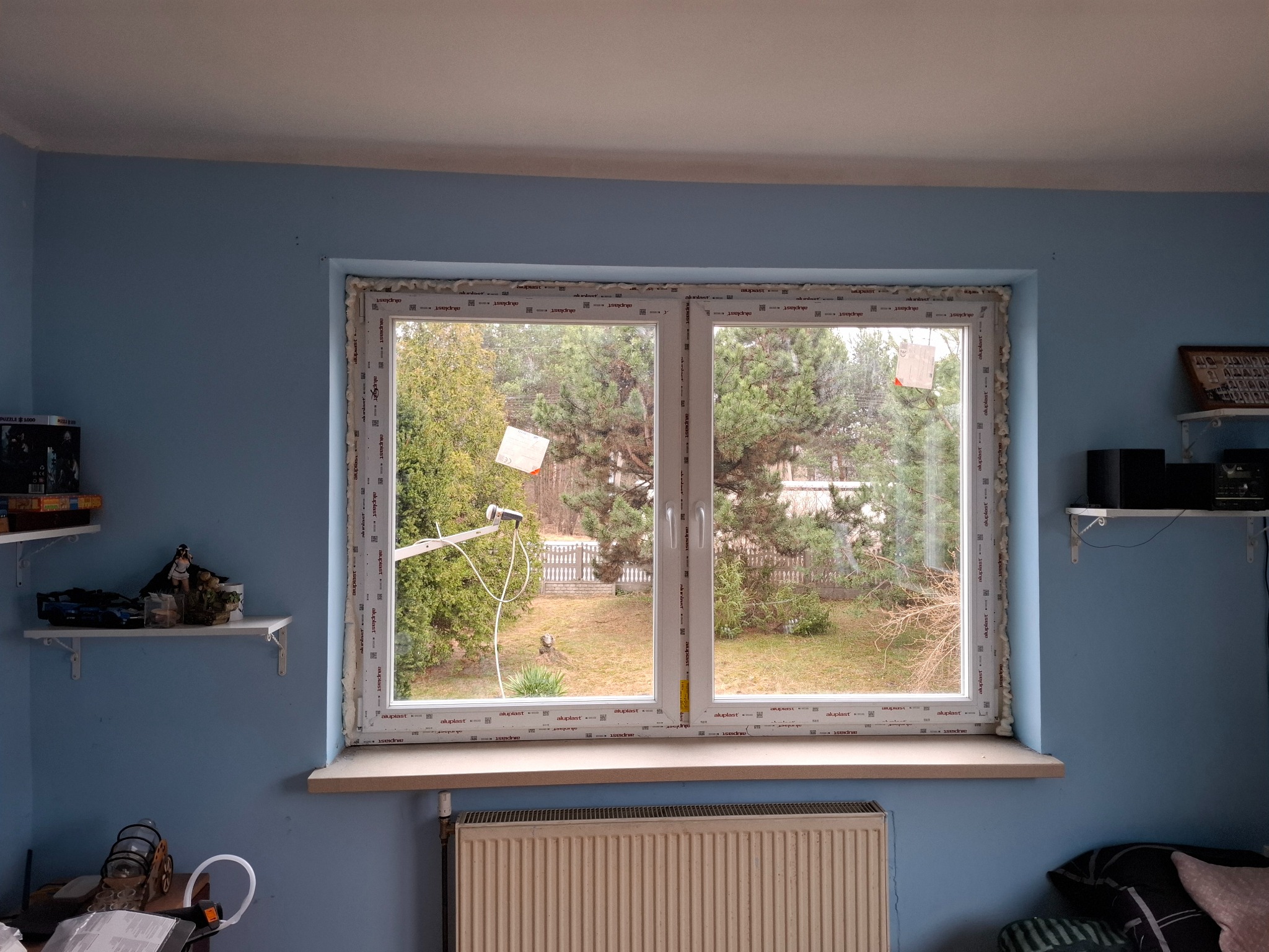 Nowe, dwuskrzydłowe okno PCV z pianką montażową, widok na ogród. Świeżo zamontowane, z widocznymi etykietami producenta na ramie. Błękitne ściany w tle.