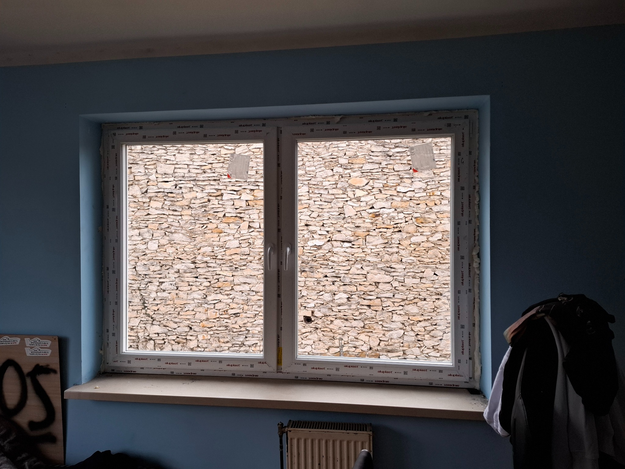 Białe okno PCV z widokiem na kamienną ścianę. Widoczne ślady montażu, niebieskie ściany w tle i parapet. Okno dwuskrzydłowe, nierozwierne. Aluplast.
