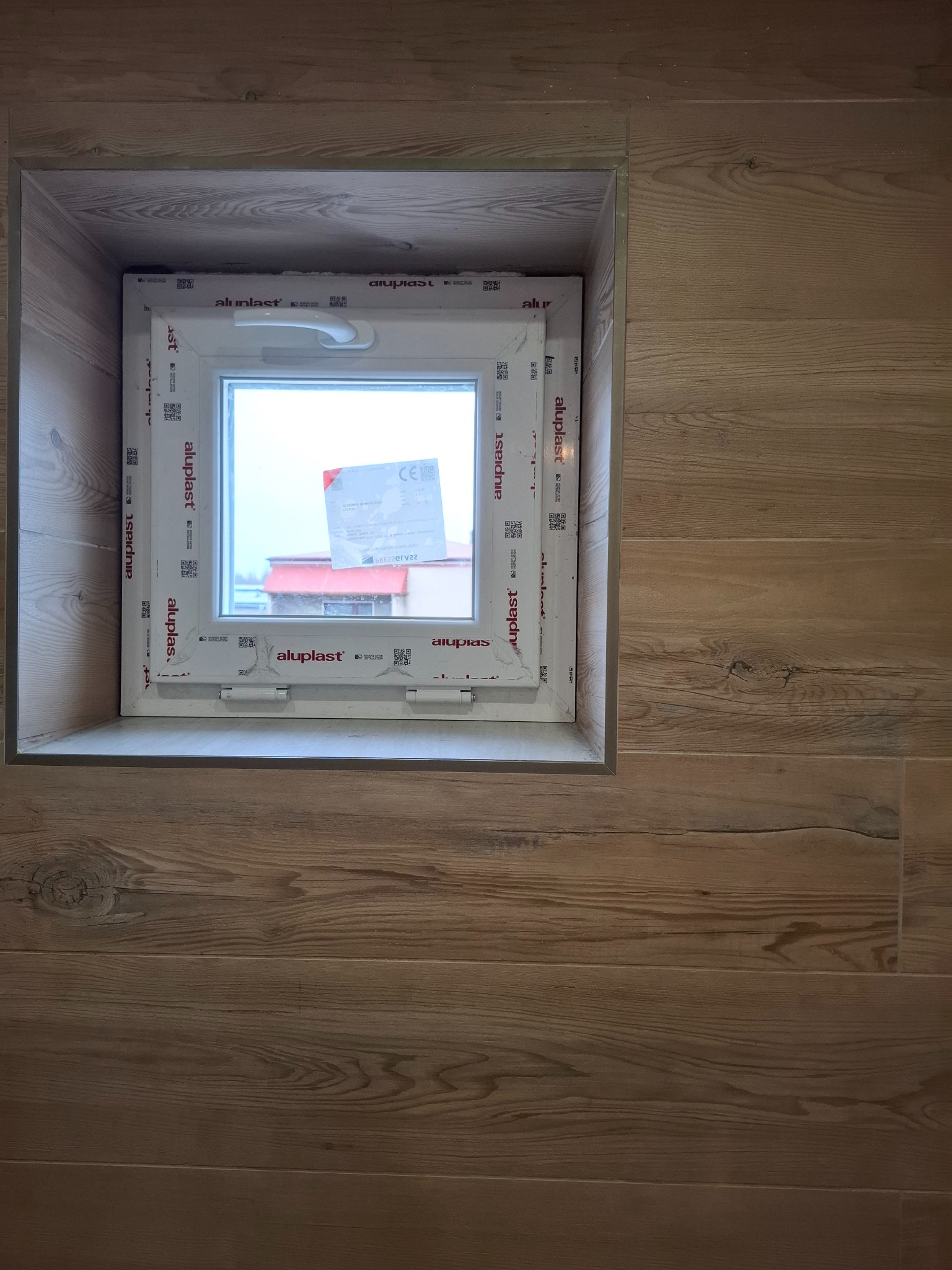 Białe okno PCV marki Aluplast, wbudowane w ścianę wyłożoną panelami imitującymi drewno, widok z bliska, z naklejkami informacyjnymi na szybie.