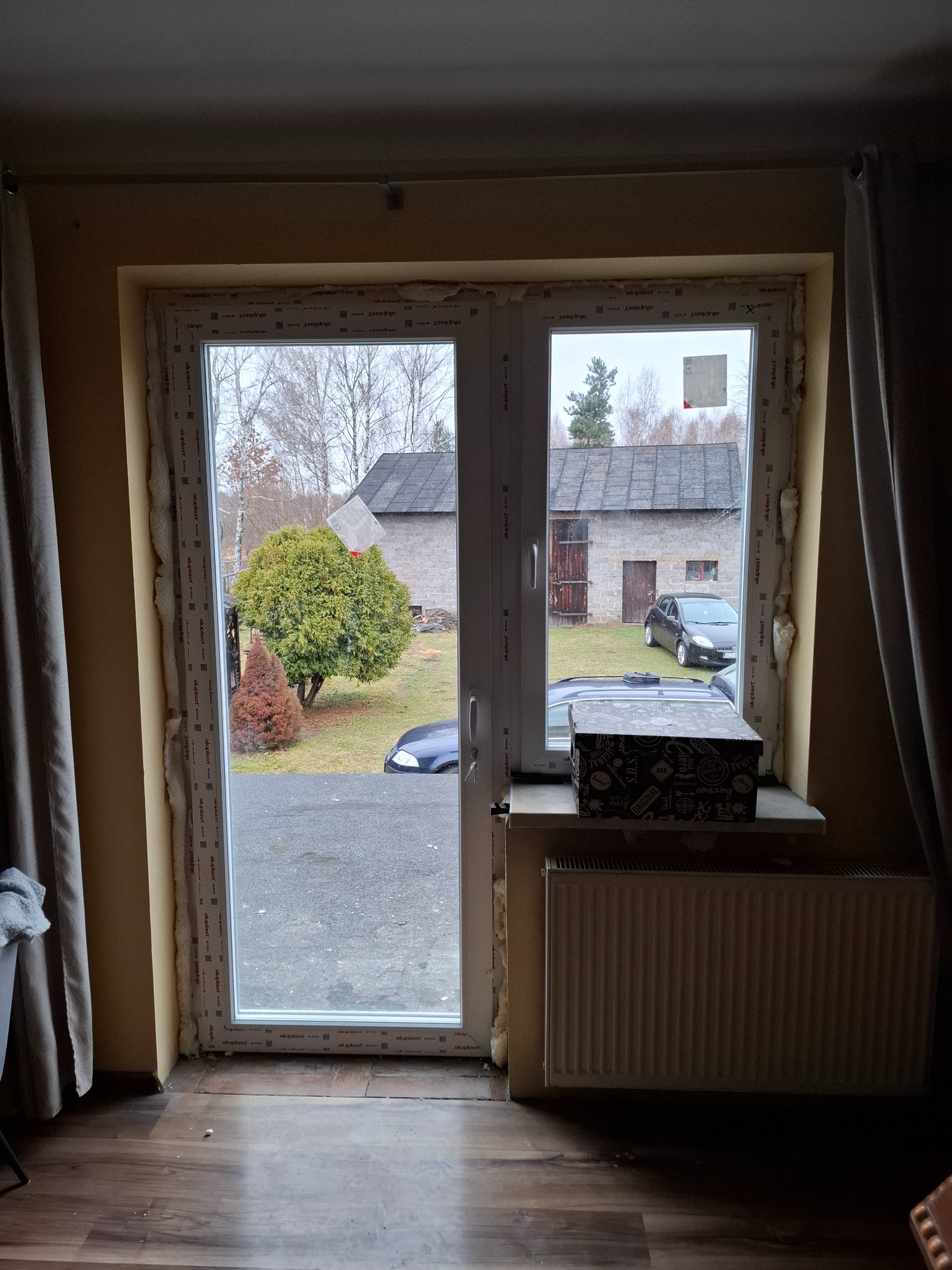 Białe okno PCV z pianką montażową, widok na podwórko z zielenią i budynkiem gospodarczym. Na parapecie pudełko, a pod nim grzejnik.