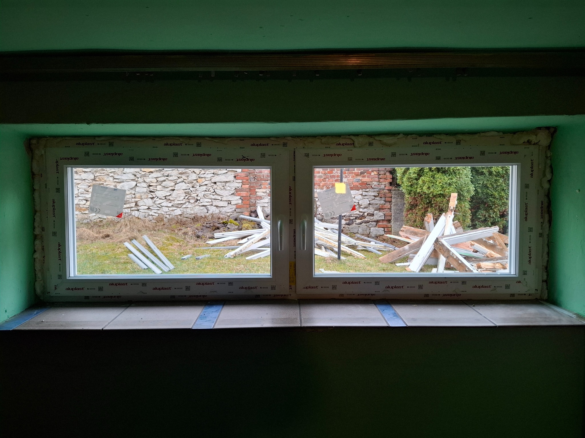 Nowo zamontowane okno PCV Aluplast z widoczną pianką montażową i elementami wykończeniowymi na zewnątrz, z fragmentem muru w tle.