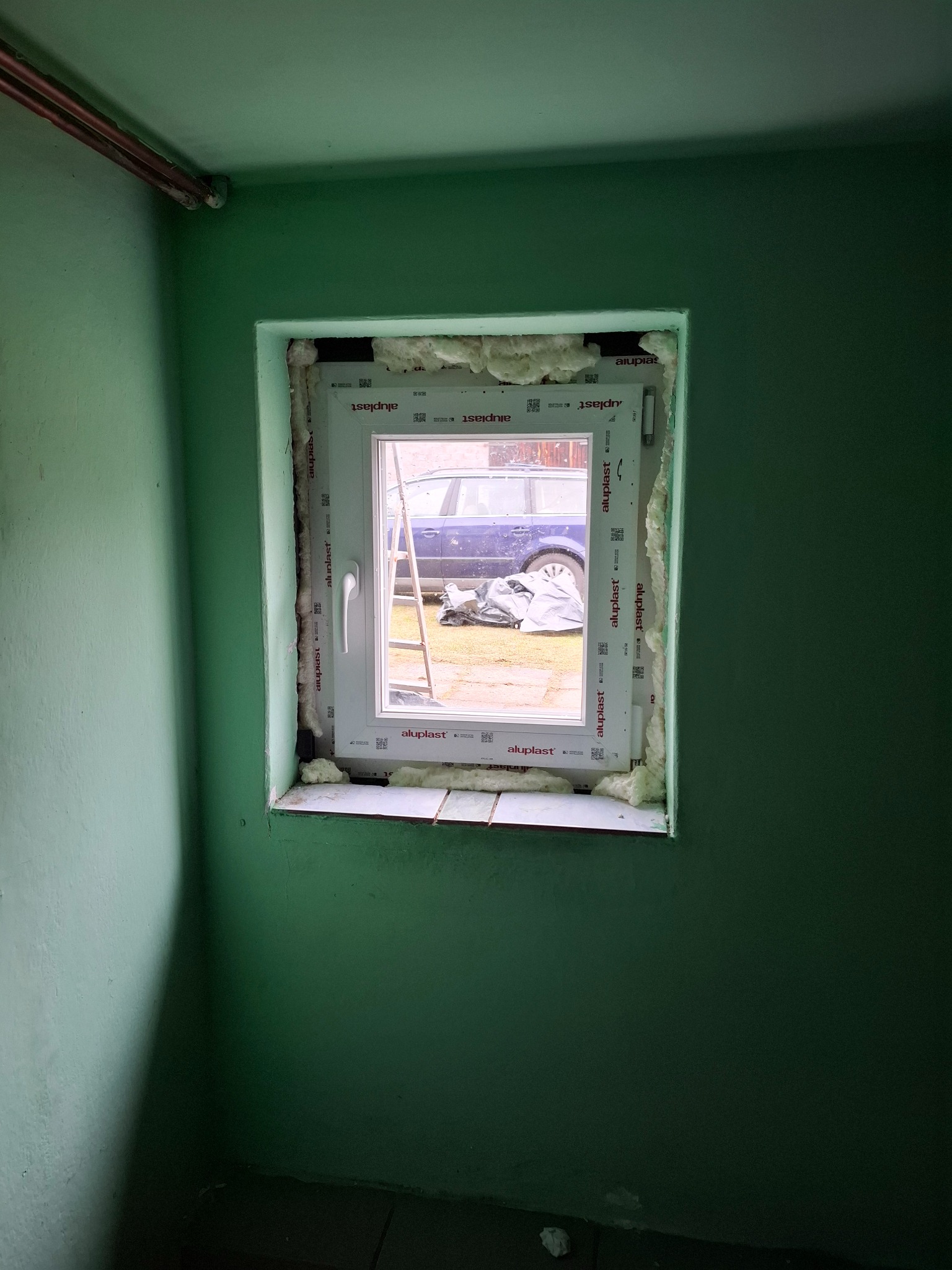 Montaż okna PCV marki Aluplast z pianką montażową, widok z wnętrza pomieszczenia o zielonych ścianach na zewnątrz, gdzie stoi drabina i zaparkowany samochód.