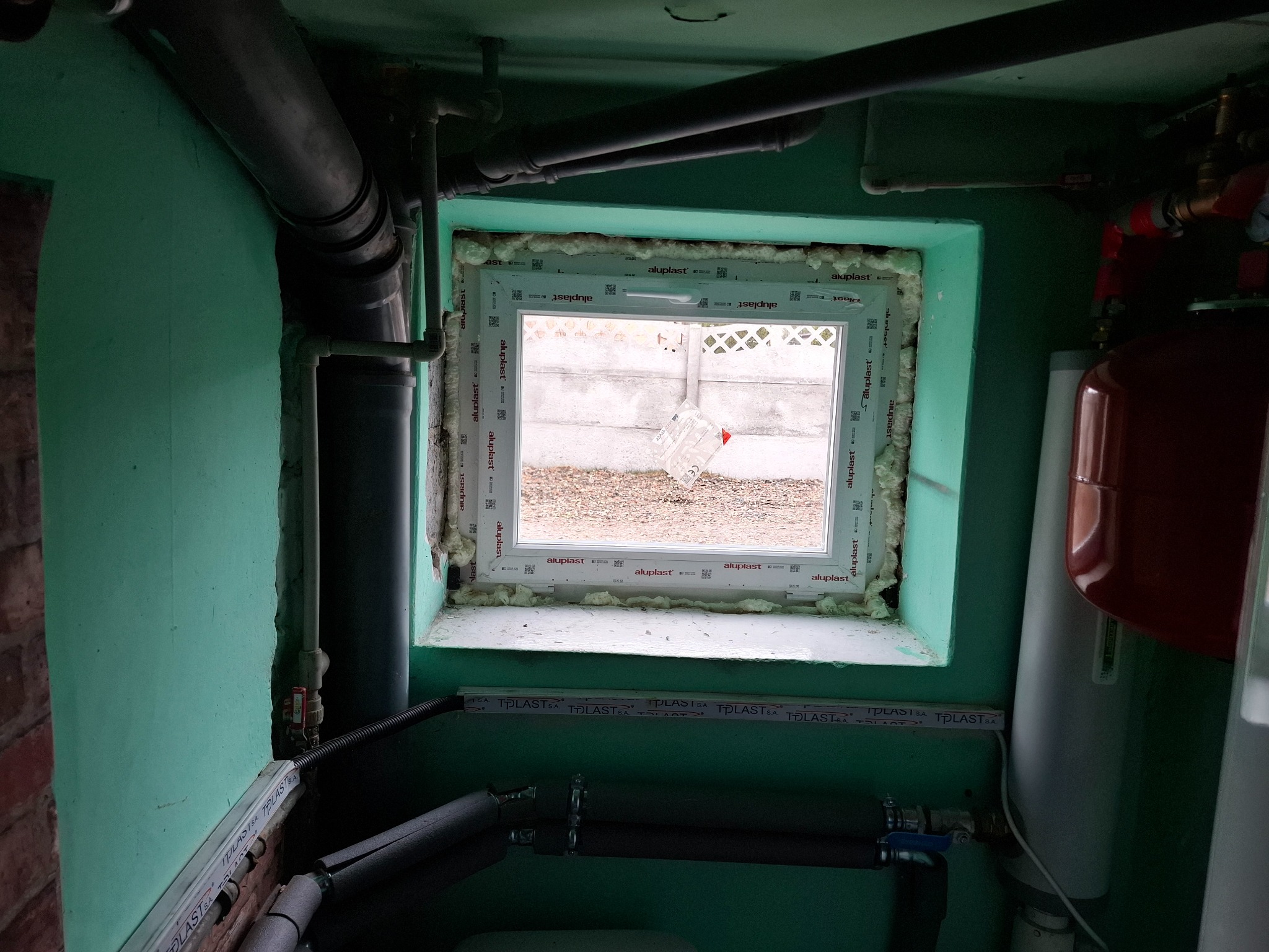 Niewielkie okno PCV Aluplast w zielonym, zagraconym pomieszczeniu, widoczne rury i instalacje. Okno uszczelnione pianką montażową, z widokiem na zewnątrz.