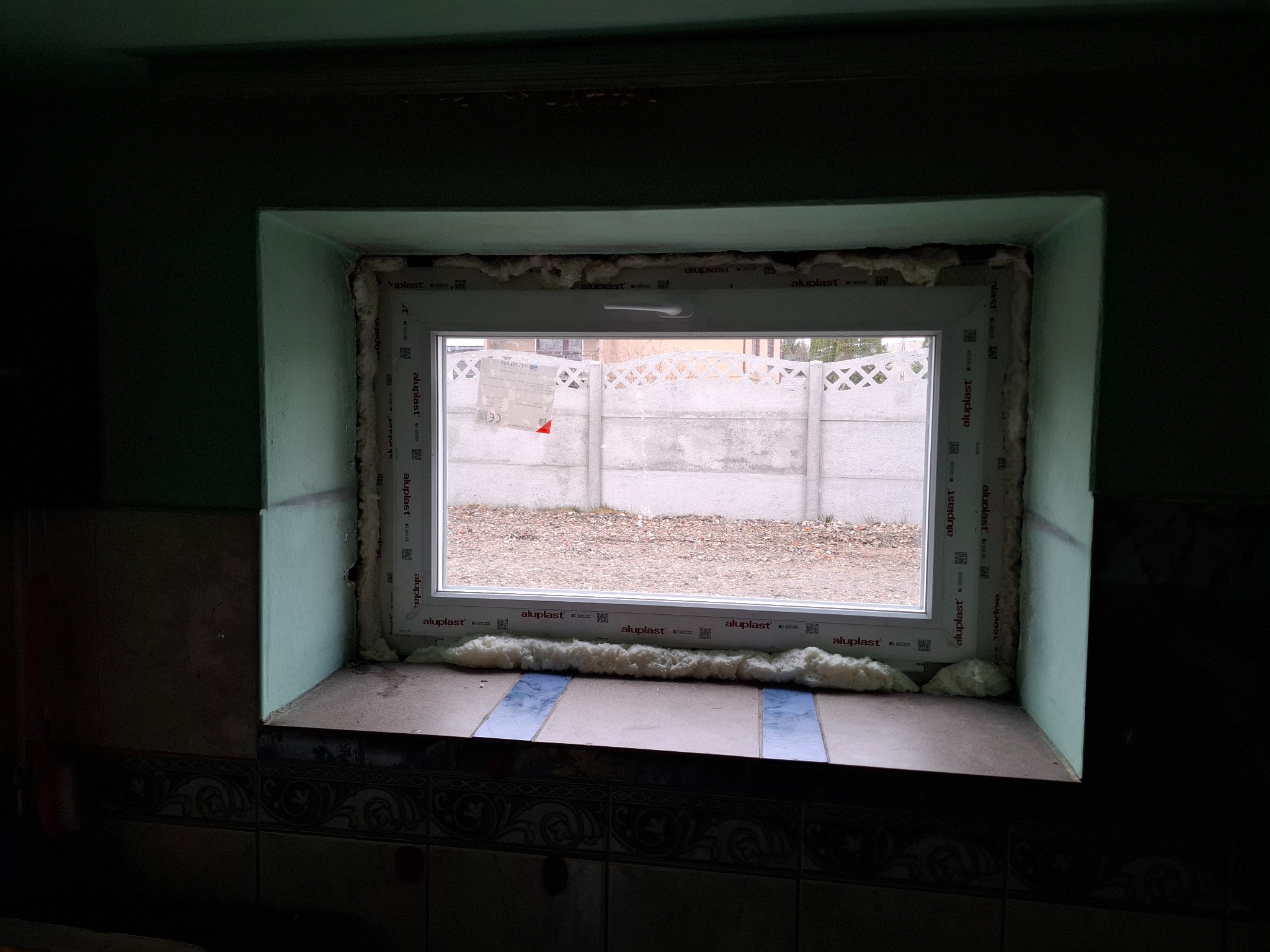 Nowe okno PCV Aluplast z białą ramą, zamontowane w ościeżnicy, uszczelnione pianką montażową. Widok na zewnątrz przez szybę na betonowy płot i ziemię.