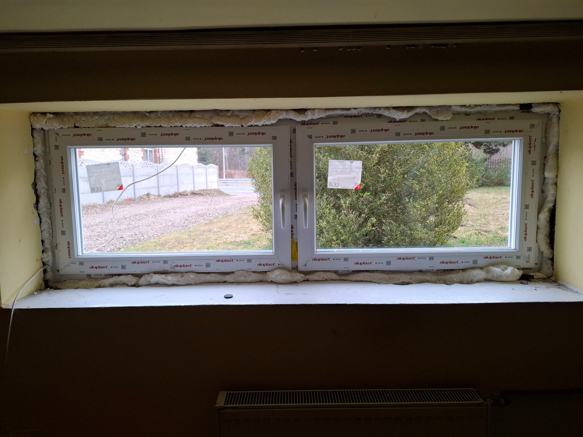 Montaż okna PCV: biała rama okienna Aluplast z pianką montażową, widok na ogród i betonowe ogrodzenie. Wnętrze w trakcie wykończenia, grzejnik pod parapetem.