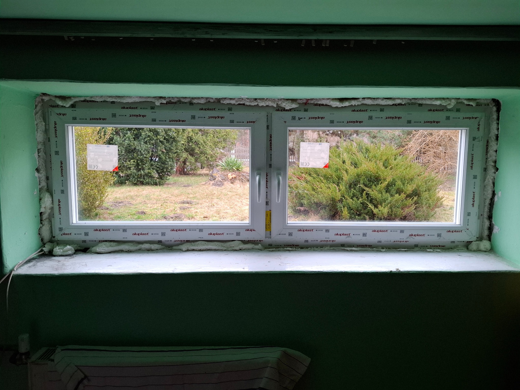 Białe okno PCV marki Aluplast, z widocznymi pianką montażową i naklejkami. Widok na zewnątrz, na zieleń. Ściana pomalowana na zielono, parapet biały.