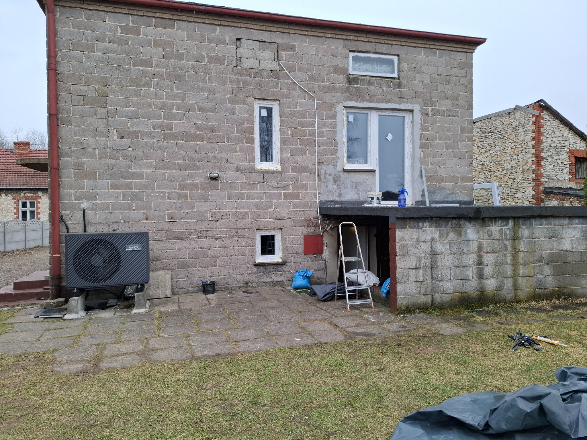 Budynek z szarego pustaka z nowymi oknami PCV w trakcie montażu, widoczna drabina i akcesoria montażowe. Częściowo ocieplony budynek, widoczne okna w różnym stadium montażu.