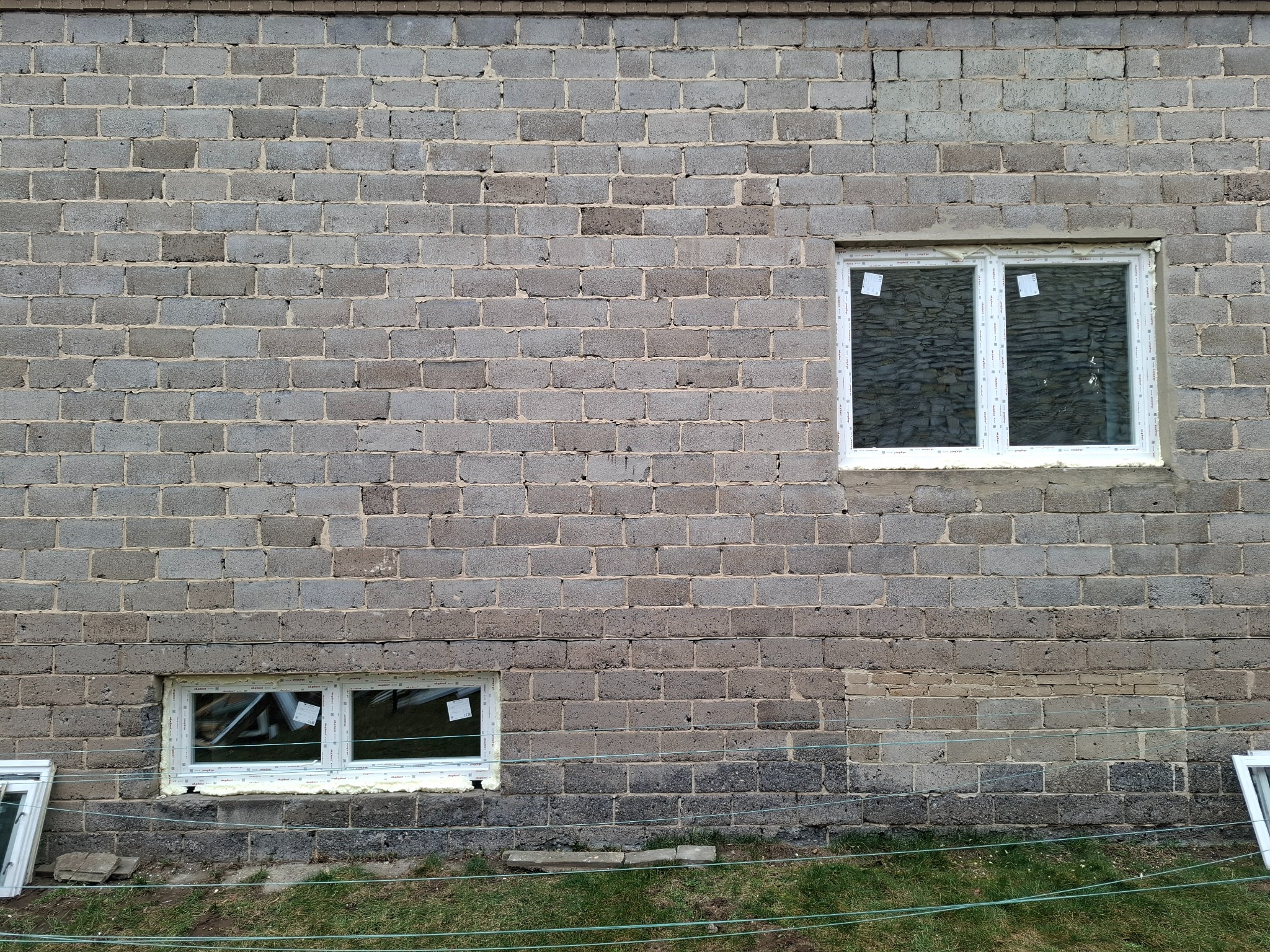 Montaż okien PCV w surowym murze z betonu komórkowego, widoczne uszczelnienia pianką montażową, dodatkowo okno obok budynku. Dwa okna o różnych rozmiarach.