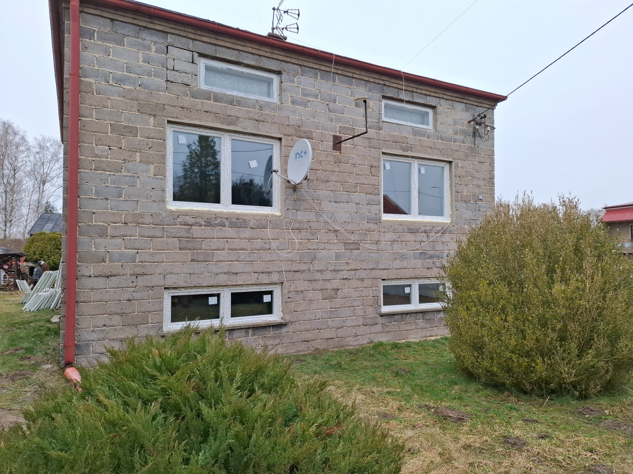 Szara bryła budynku z białymi oknami PCV. Widoczne krzewy i antena satelitarna. Częściowo otynkowana ściana. Na trawniku widoczne ślady po kretach.