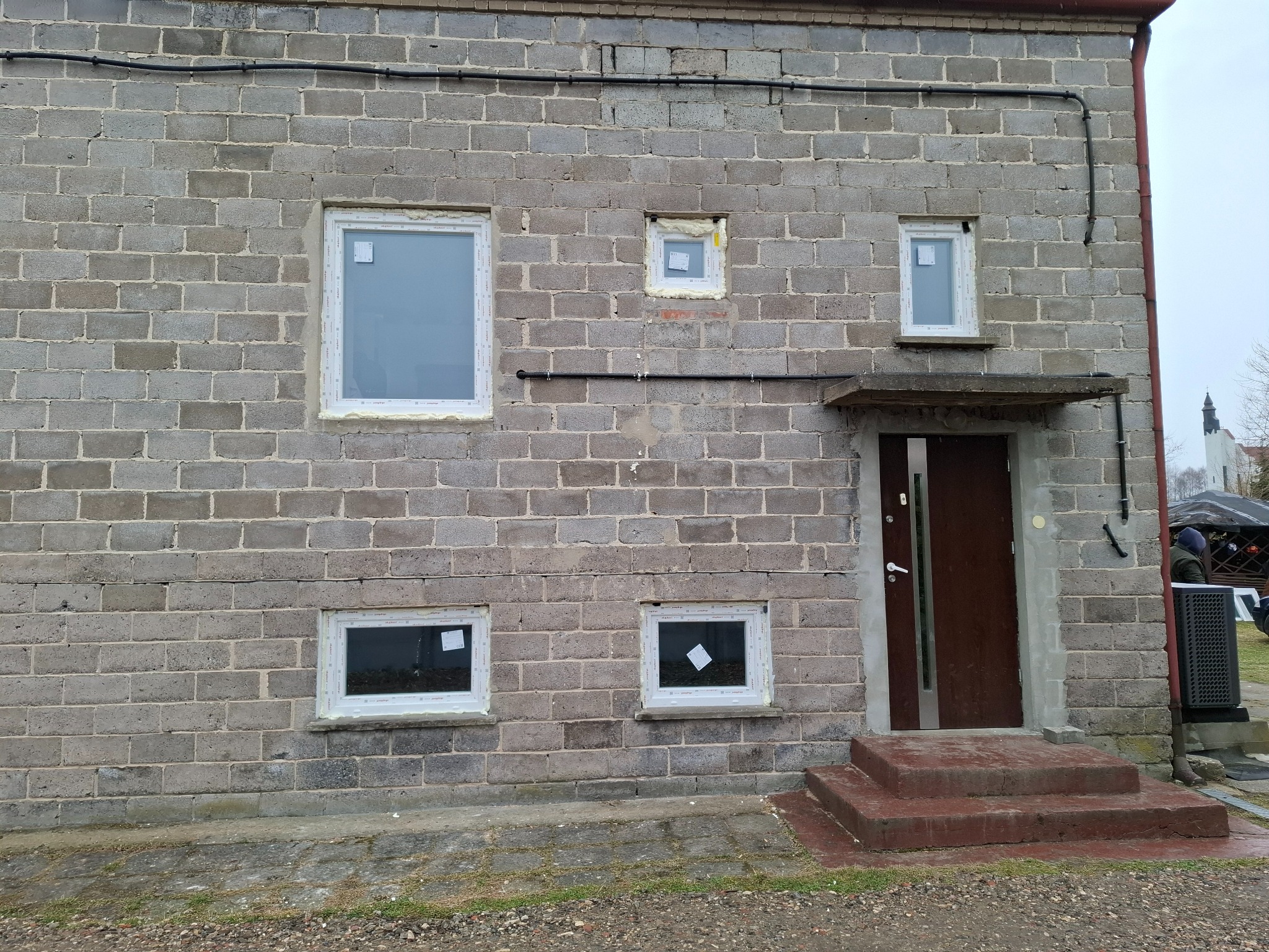 Murek z szarego betonu komórkowego z zamontowanymi oknami PCV i brązowymi drzwiami wejściowymi pod betonowym daszkiem. Widoczne czarne kable na ścianie.