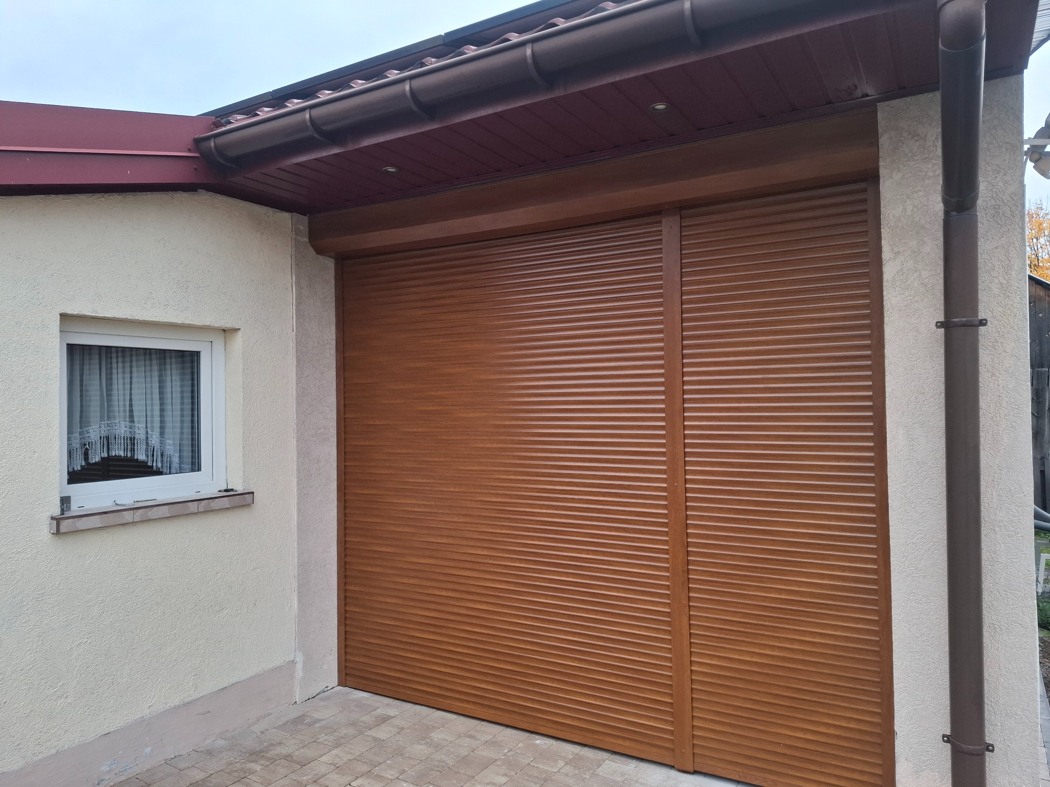 Zewnętrzna, brązowa roleta garażowa zamontowana na budynku z oknem. Widoczny fragment dachu z rynną. Elewacja budynku w jasnym kolorze.
