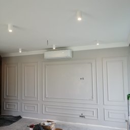 Zakład Remontowo-Budowlany Czesław Kamiński - Pokój w trakcie remontu z ozdobnymi panelami ściennymi w stylu klasycznym, widoczne narzędzia i materiały budowlane na podłodze, klimatyzator na ścianie, sufit z oświetleniem punktowym.