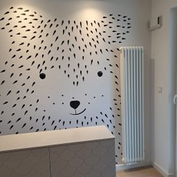 Zakład Remontowo-Budowlany Czesław Kamiński - Ściana pokoju dziecięcego z naklejką w kształcie niedźwiedzia, biała komoda z szarymi drzwiczkami o wzorze rybiej łuski i grzejnik żeberkowy.