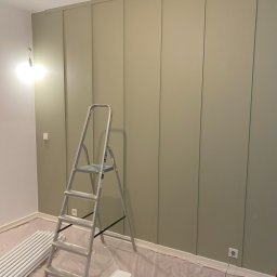 Zakład Remontowo-Budowlany Czesław Kamiński - Ściana z panelami pomalowana na oliwkowy kolor, zabezpieczona folią podłoga, drabina aluminiowa, rury grzejnikowe i wiadro z farbą w trakcie malowania.