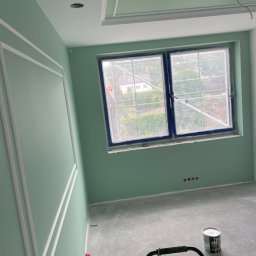 Zakład Remontowo-Budowlany Czesław Kamiński - Pokój po świeżym pomalowaniu na pistacjowy kolor z białymi sztukateriami na ścianie i suficie. Widoczne okno z niebieską ramą, zabezpieczone folią, oraz puszka z farbą i urządzenie czyszczące...