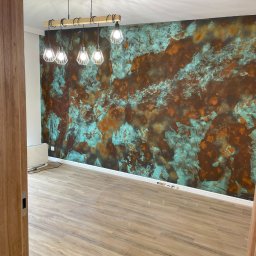 Zakład Remontowo-Budowlany Czesław Kamiński - Pusty pokój z dekoracyjną ścianą w odcieniach miedzi i turkusu, oświetlony nowoczesną lampą wiszącą z drewnianym elementem, panele podłogowe imitujące drewno, widok zza otwartych drzwi.