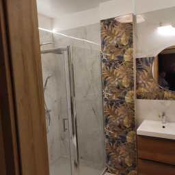 Zakład Remontowo-Budowlany Czesław Kamiński - Nowoczesna łazienka z kabiną prysznicową, umywalką z szafką z drewna oraz płytkami z motywem roślinnym na ścianie, widok zza drewnianej ościeżnicy drzwi.