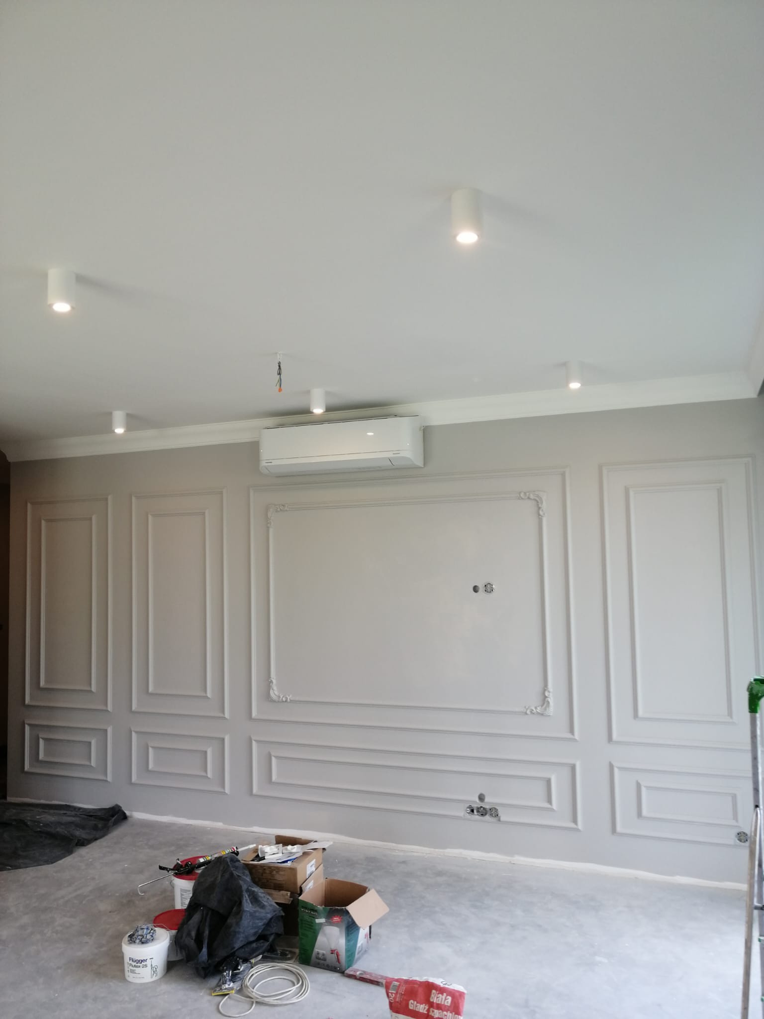 Pokój w trakcie remontu z ozdobnymi panelami ściennymi w stylu klasycznym, widoczne narzędzia i materiały budowlane na podłodze, klimatyzator na ścianie, sufit z oświetleniem punktowym.