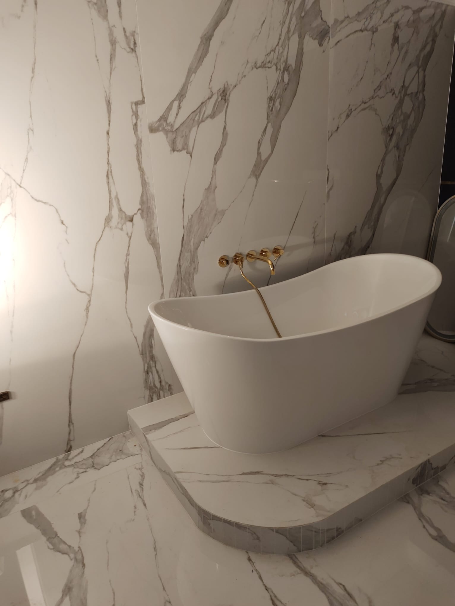 Elegancka, wolnostojąca wanna w łazience wykończonej marmurowymi płytkami z subtelnymi szarymi żyłkami, bateria w kolorze szczotkowanego złota.
