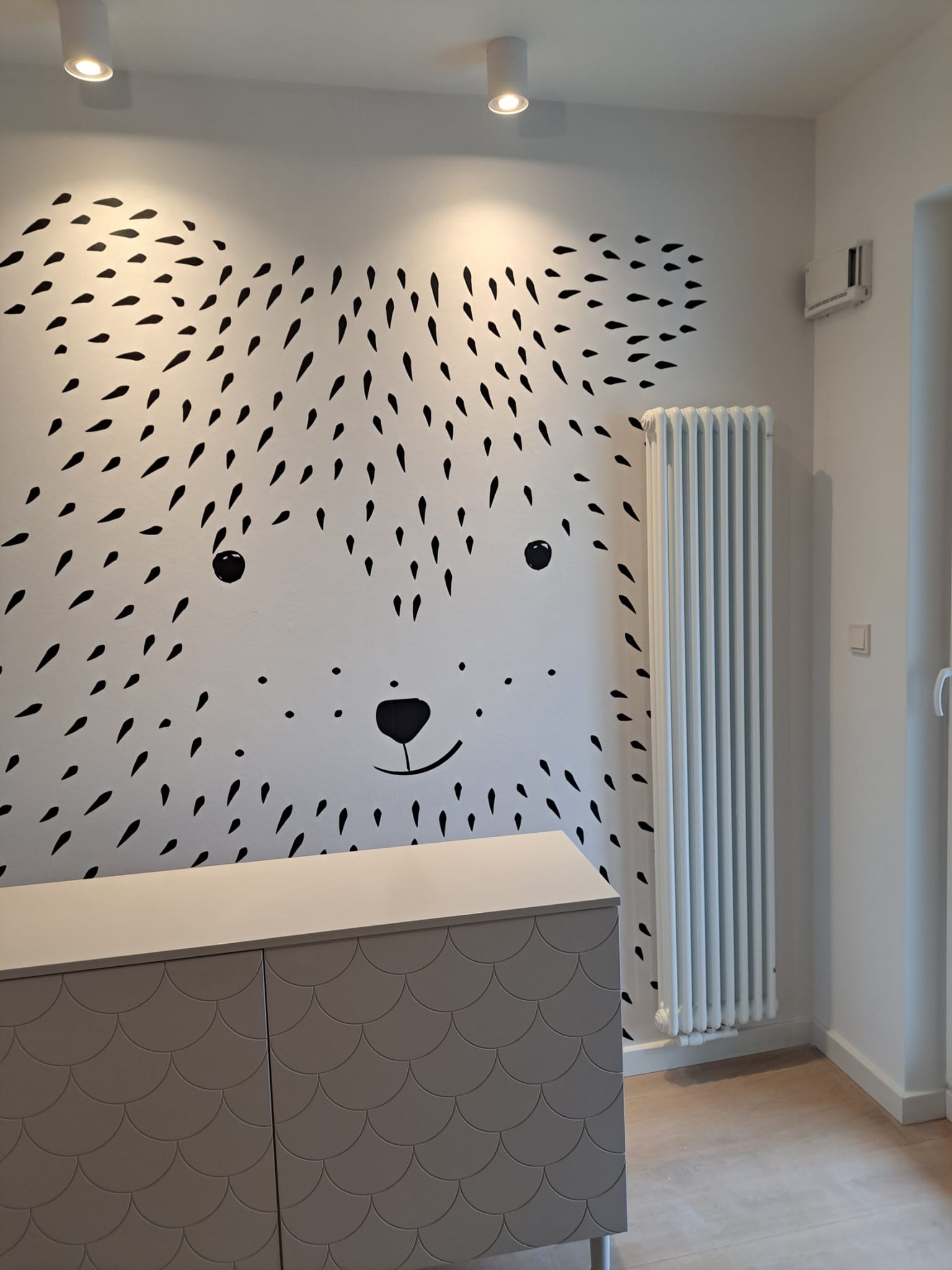 Ściana pokoju dziecięcego z naklejką w kształcie niedźwiedzia, biała komoda z szarymi drzwiczkami o wzorze rybiej łuski i grzejnik żeberkowy.