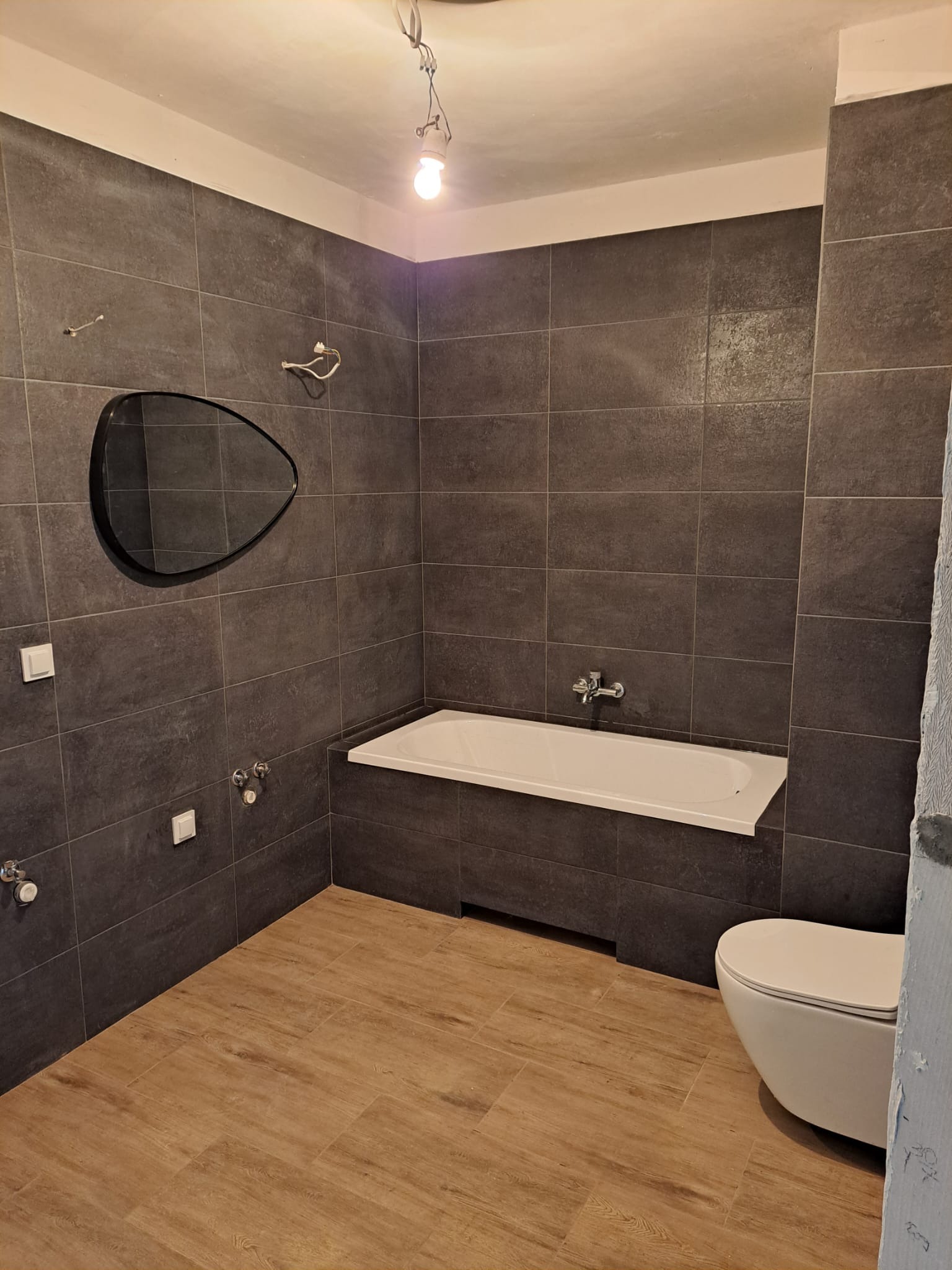 Nowoczesna łazienka z ciemnoszarymi płytkami, drewnopodobną podłogą, asymetrycznym lustrem i wiszącą żarówką bez klosza.