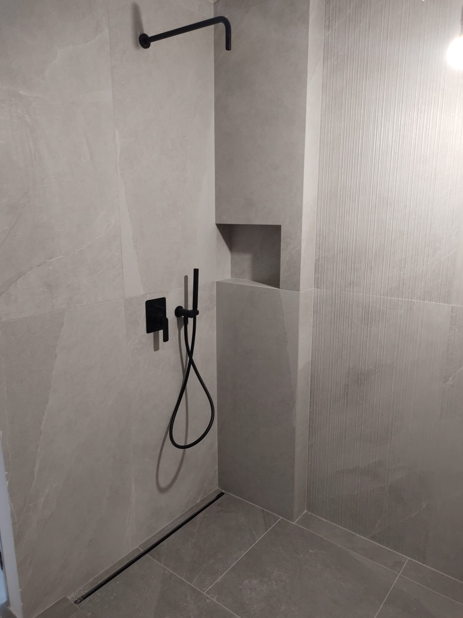 Nowoczesna kabina prysznicowa z szarymi płytkami imitującymi kamień, czarna armatura: deszczownica, bateria podtynkowa i wąż prysznicowy, odpływ liniowy w podłodze, półka w ścianie.
