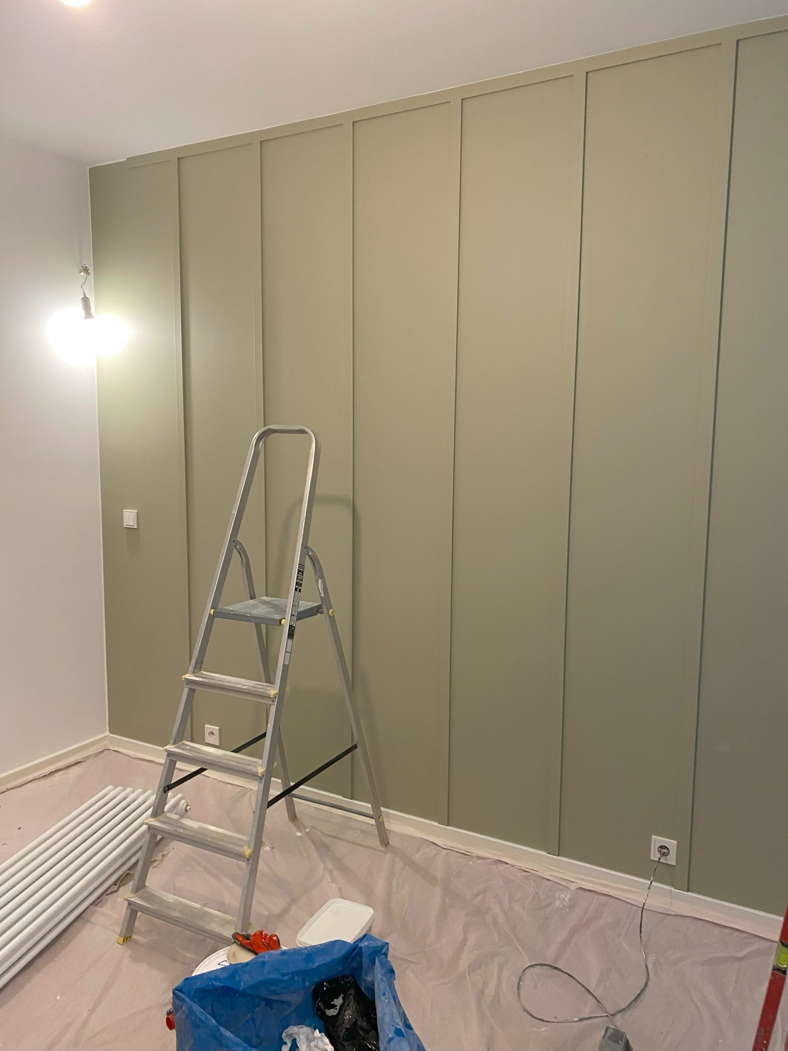 Ściana z panelami pomalowana na oliwkowy kolor, zabezpieczona folią podłoga, drabina aluminiowa, rury grzejnikowe i wiadro z farbą w trakcie malowania.
