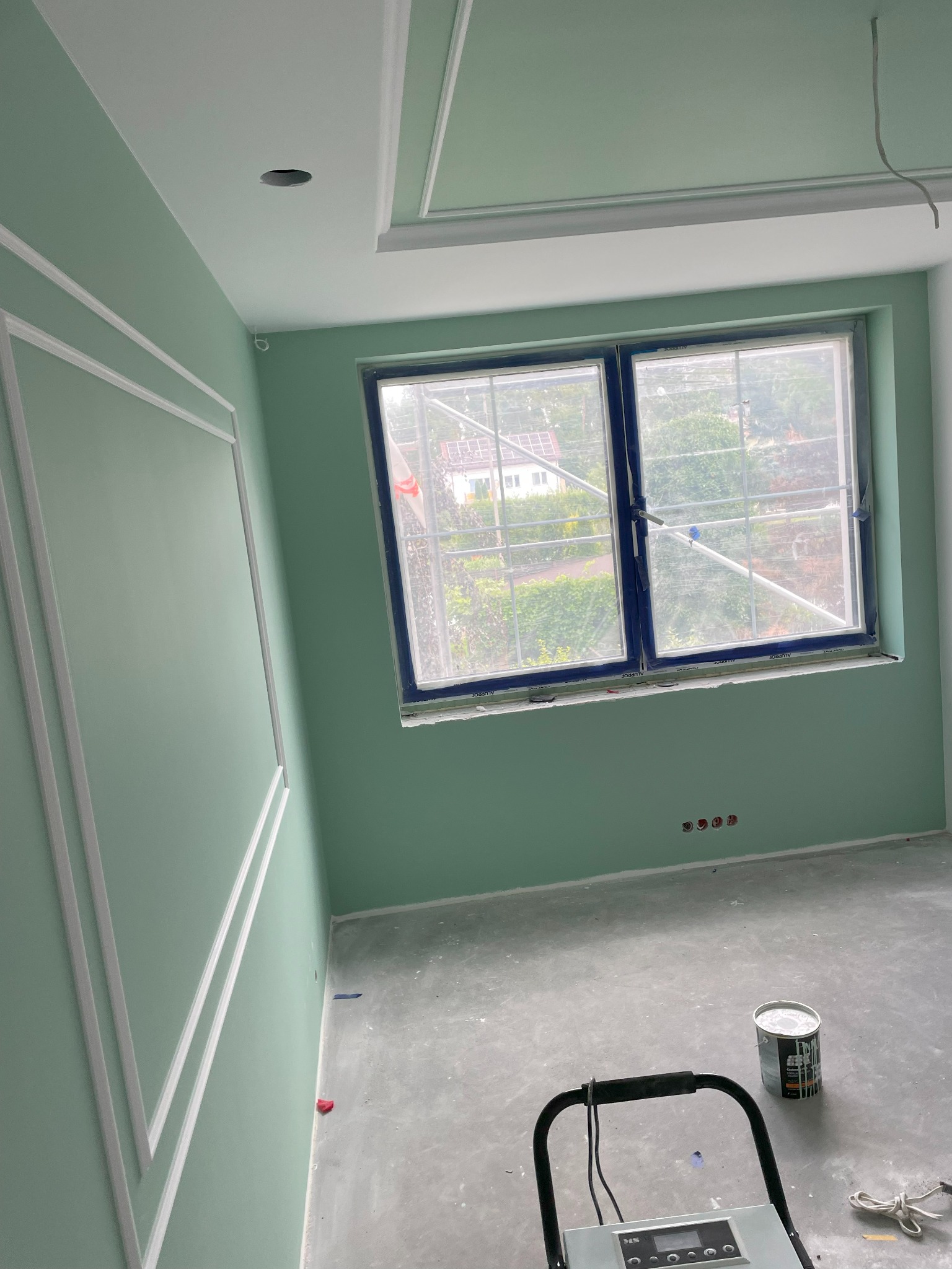 Pokój po świeżym pomalowaniu na pistacjowy kolor z białymi sztukateriami na ścianie i suficie. Widoczne okno z niebieską ramą, zabezpieczone folią, oraz puszka z farbą i urządzenie czyszczące...