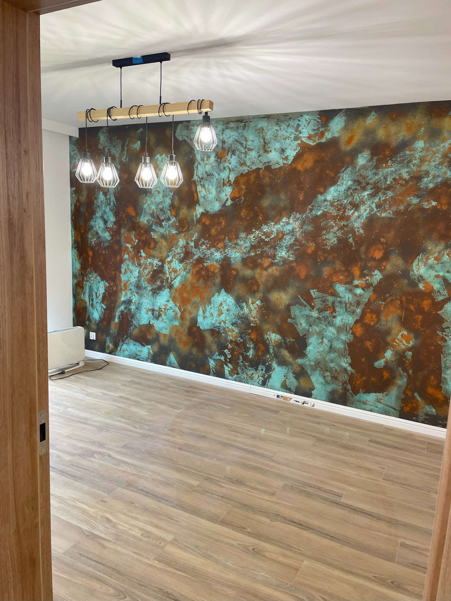 Pusty pokój z dekoracyjną ścianą w odcieniach miedzi i turkusu, oświetlony nowoczesną lampą wiszącą z drewnianym elementem, panele podłogowe imitujące drewno, widok zza otwartych drzwi.