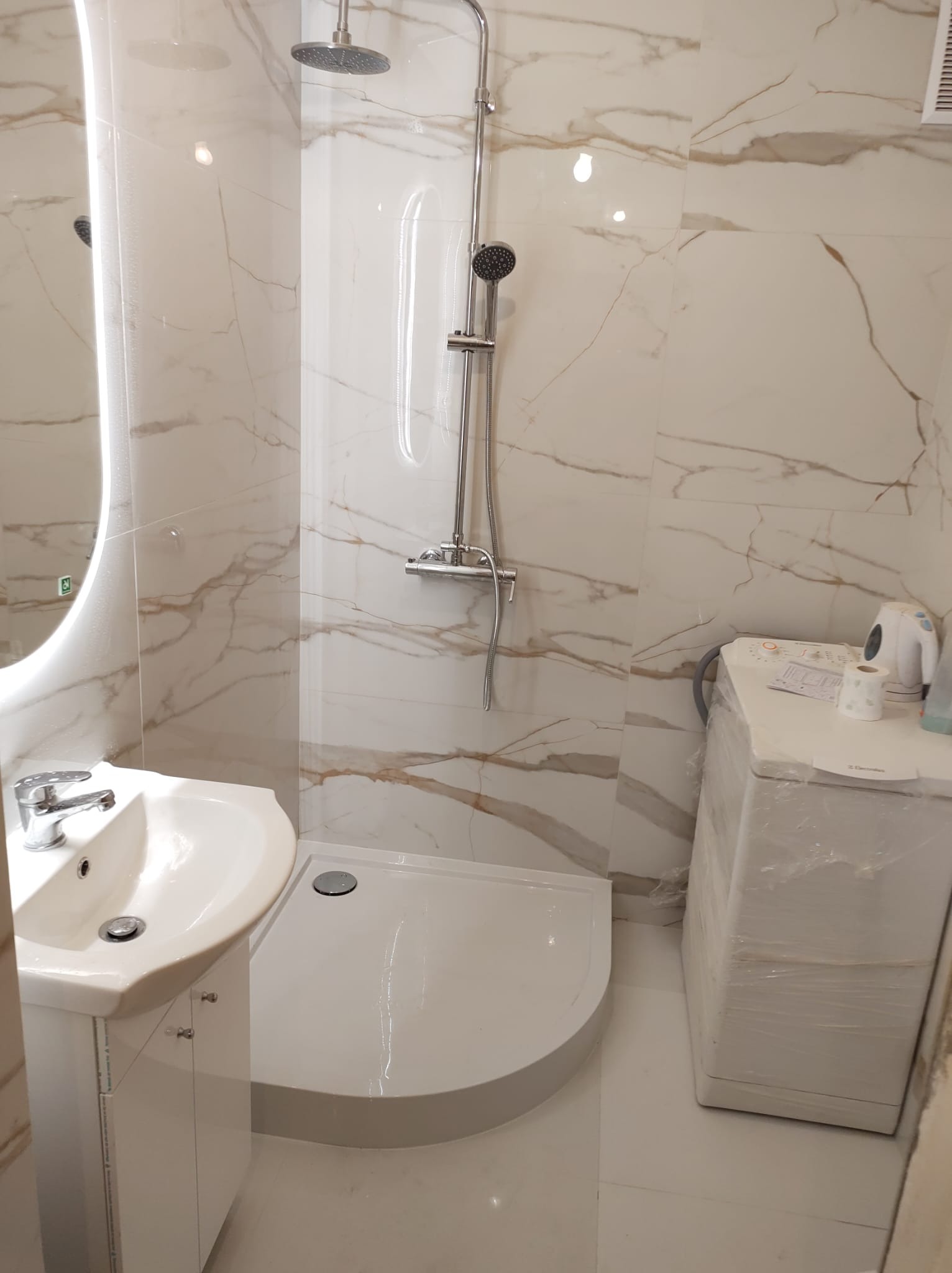 Nowoczesna łazienka z marmurowymi płytkami, kabiną prysznicową typu walk-in, umywalką z szafką i pralką owiniętą folią ochronną.