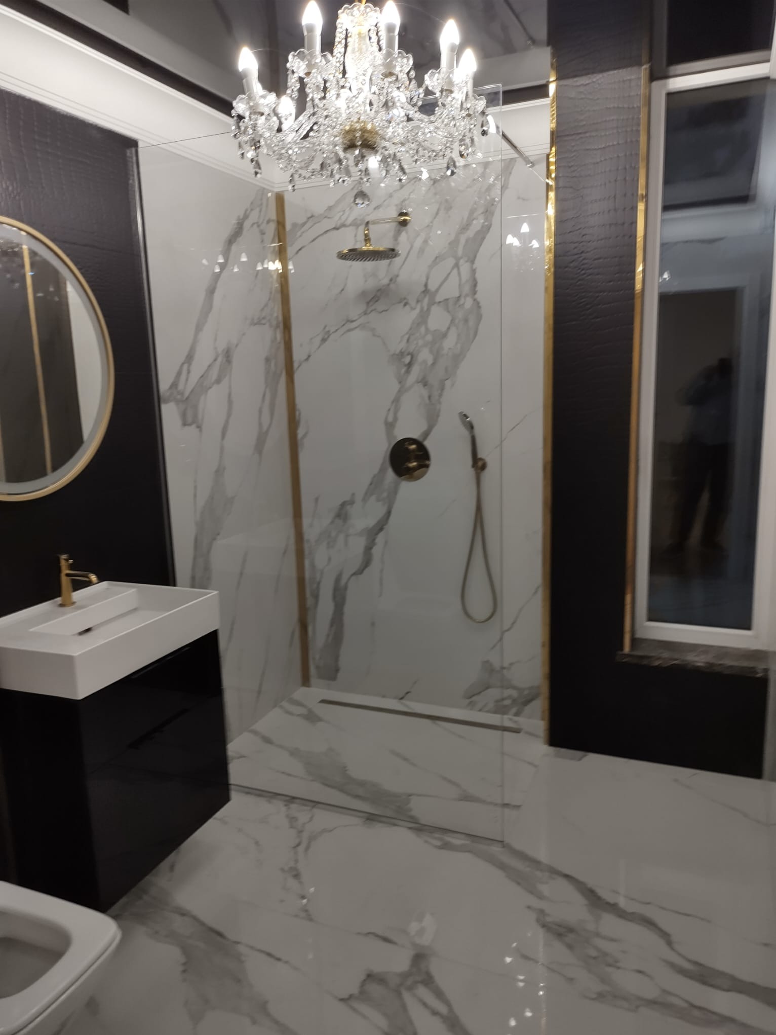 Elegancka łazienka z marmurowymi płytkami, szklanym prysznicem, złotymi akcentami i kryształowym żyrandolem, widok na umywalkę z czarną szafką i okrągłe lustro.
