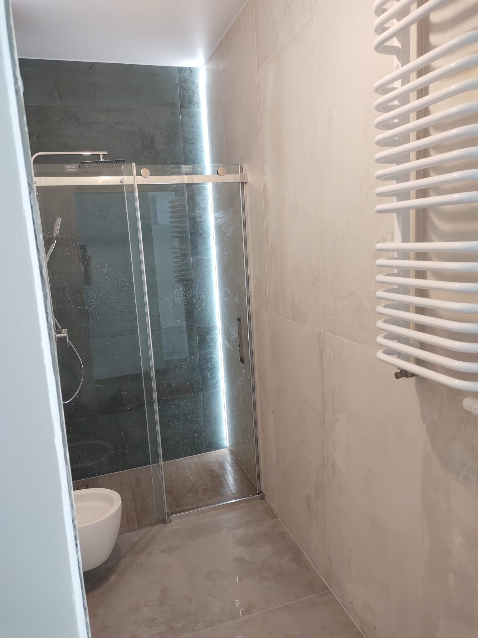 Nowoczesna łazienka z kabiną prysznicową typu walk-in, wykończona płytkami imitującymi beton, z grzejnikiem drabinkowym i podświetleniem LED.