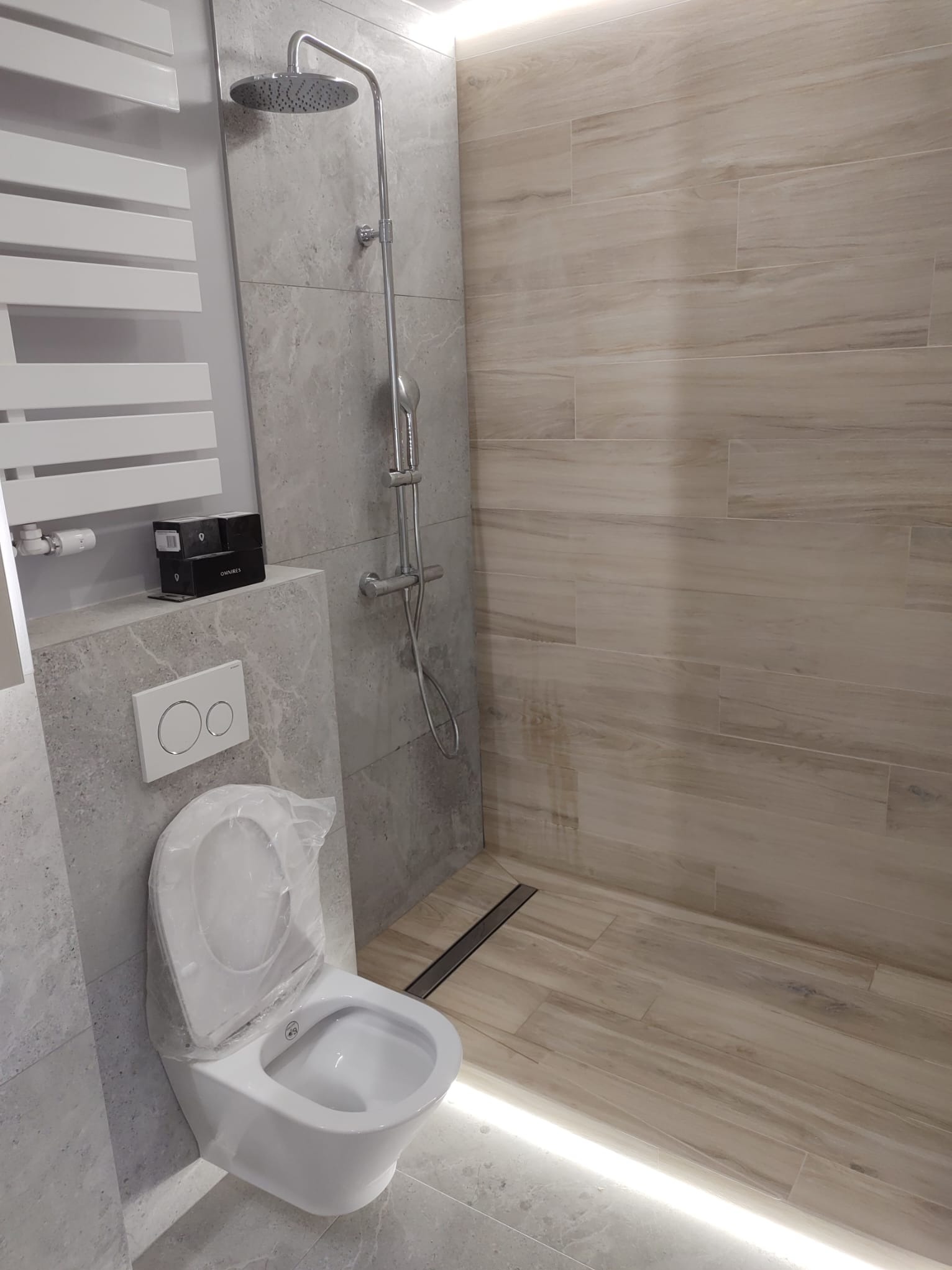 Nowoczesna łazienka z podświetleniem LED pod prysznicem, płytkami imitującymi drewno na ścianie i podłodze, białą toaletą z otwartą klapą zabezpieczoną folią oraz grzejnikiem drabinkowym i armaturą...